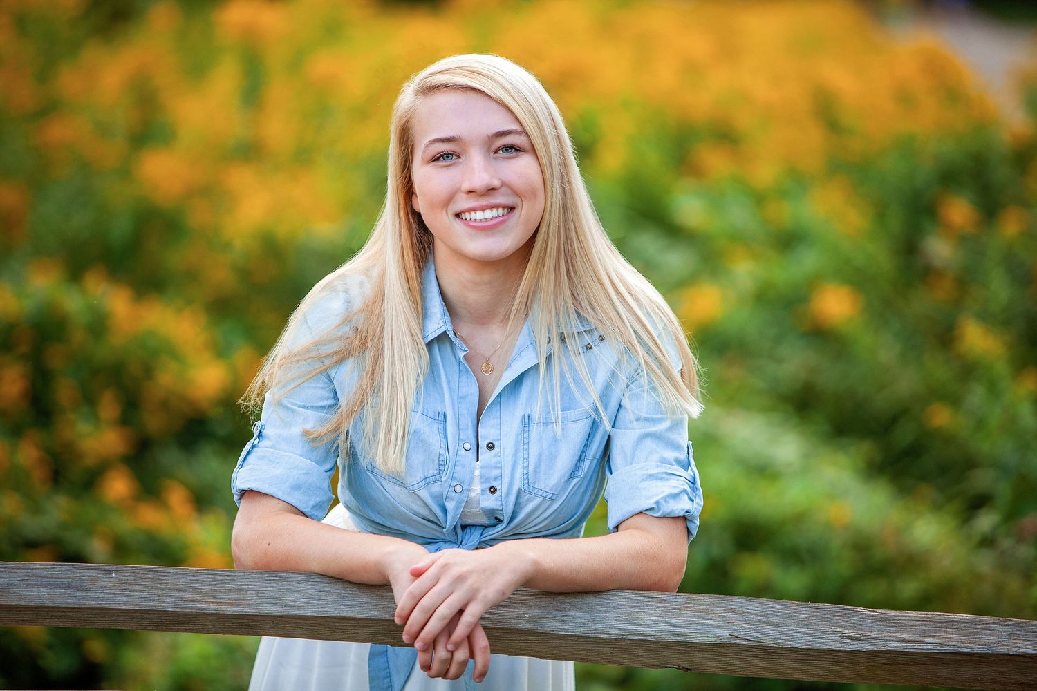 Smiling blonde teen girl leaning on a fence with flowers behind her.