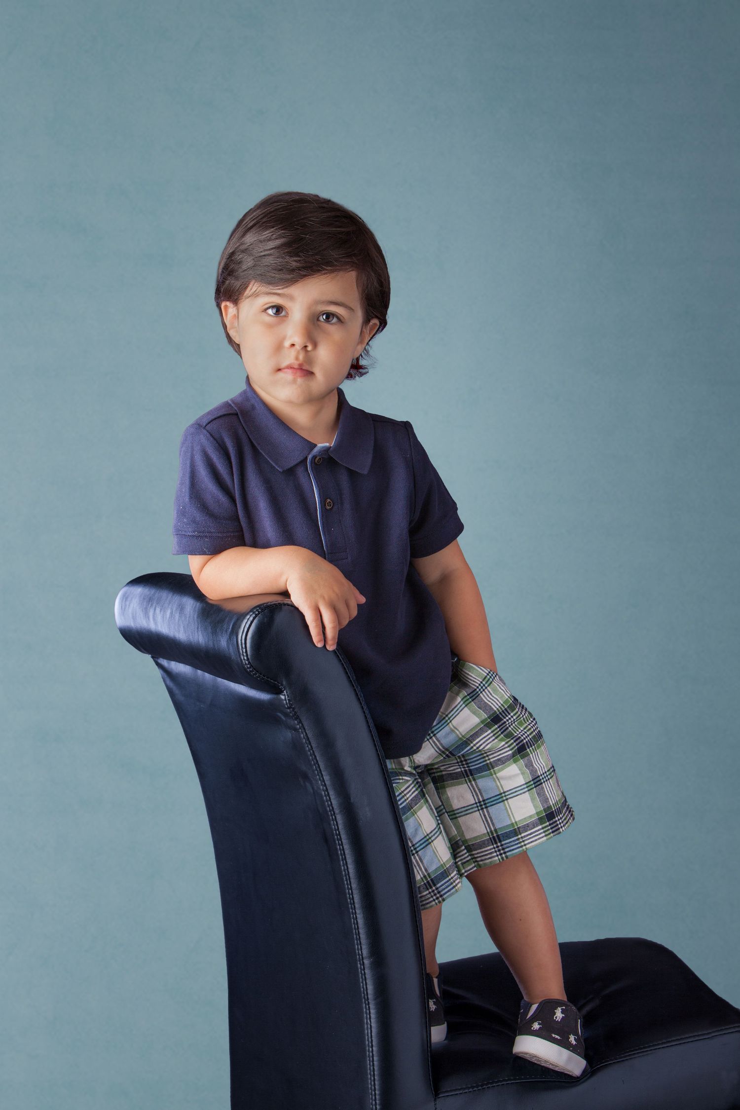 A young boy in a navy polo shirt and plaid shorts poses on a blue leather chair against a teal background.