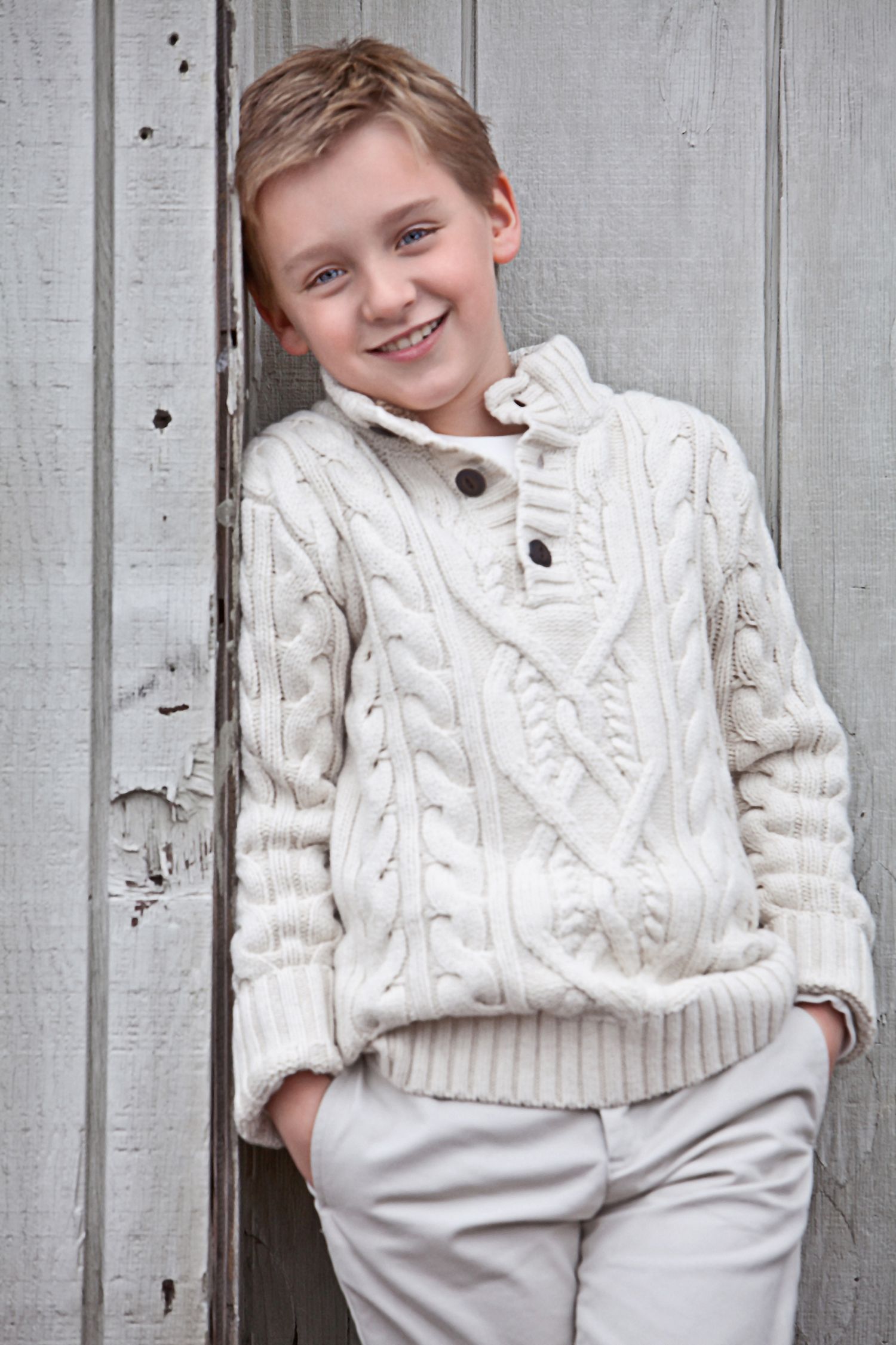 Smiling boy wearing a cream cable-knit sweater leaning against a rustic wooden wall.