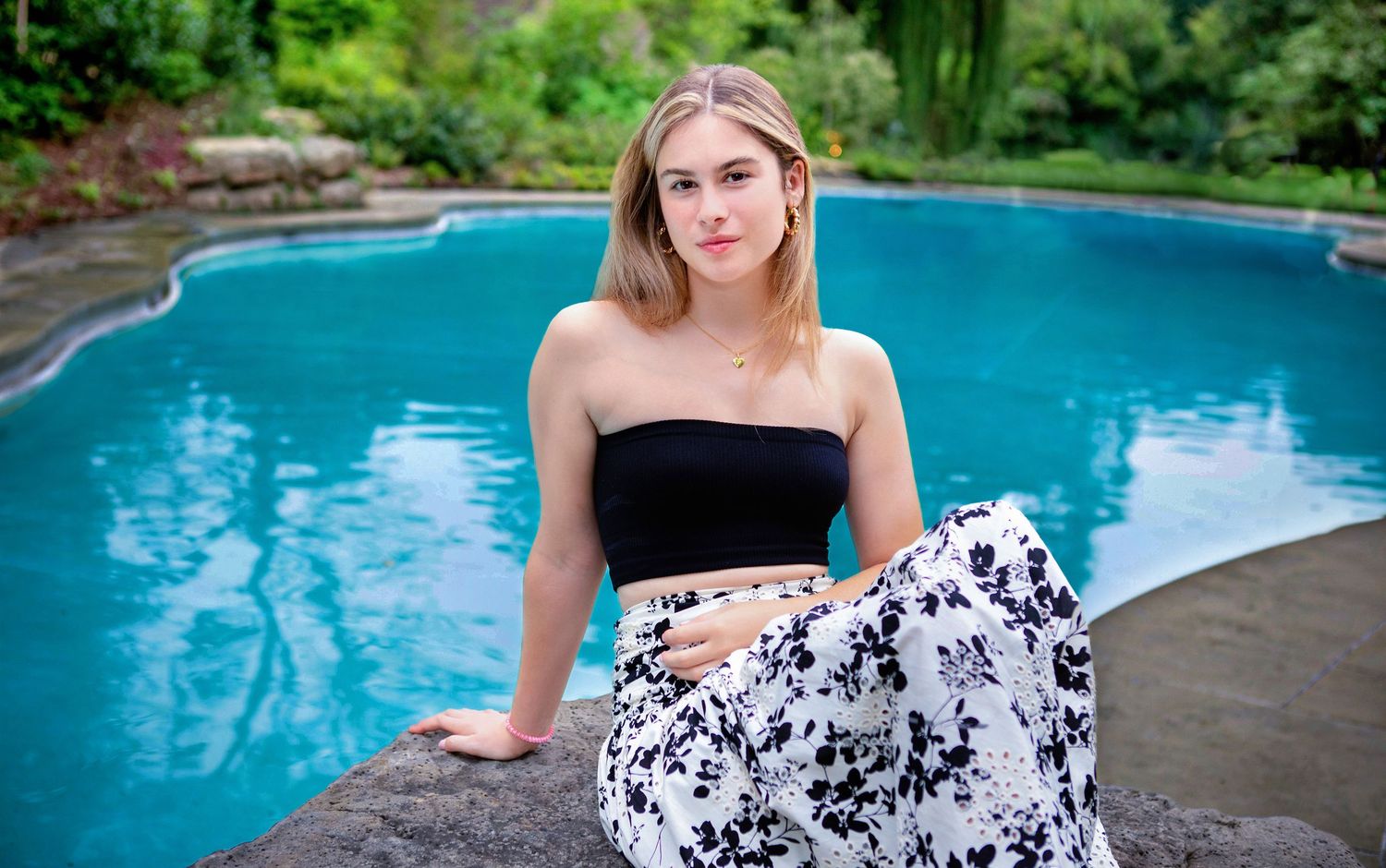 Young woman in black crop top and floral skirt posing by a blue swimming pool surrounded by lush greenery.