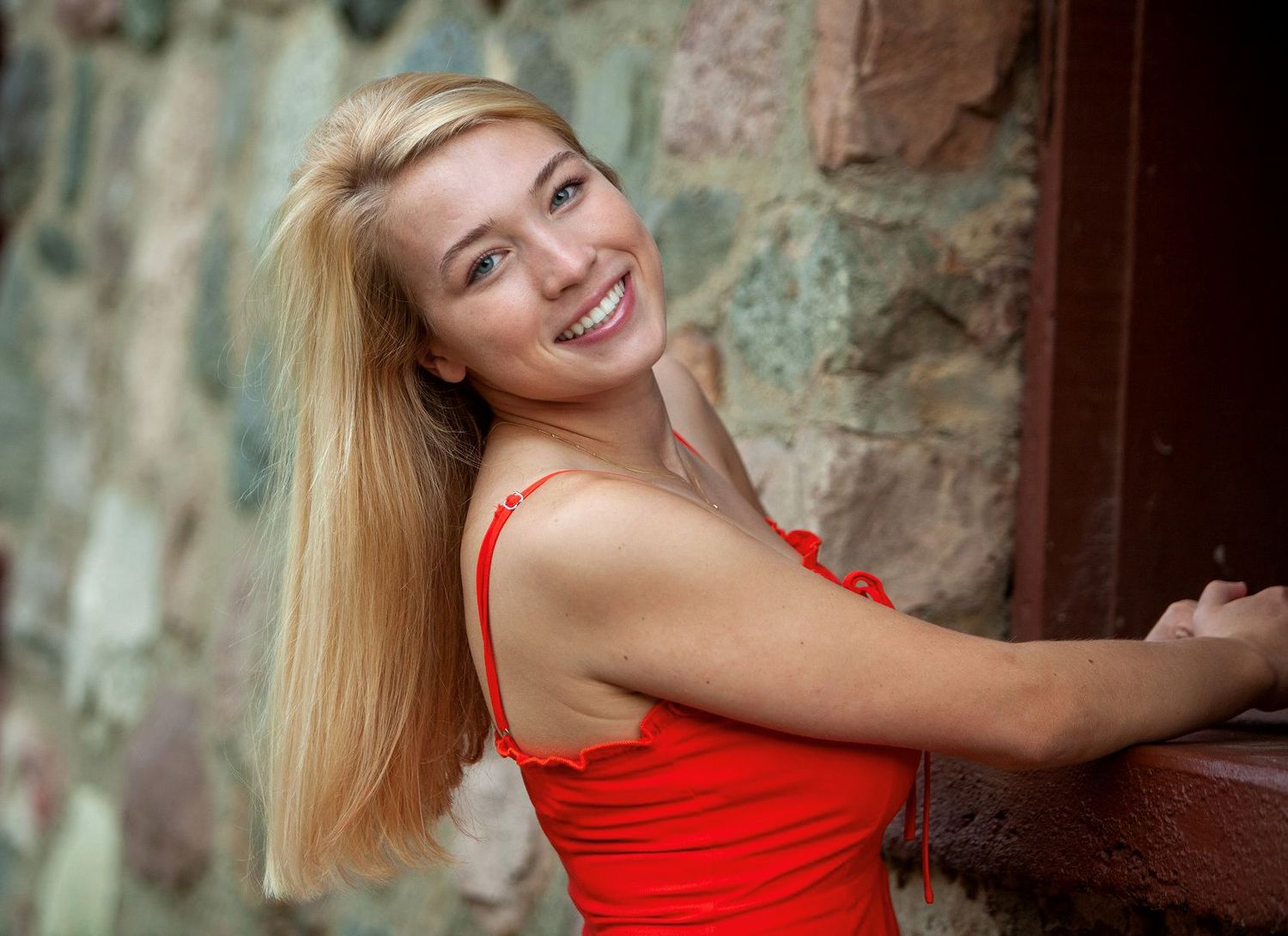 Smiling blonde young woman in red top posing by stone wall in outdoor portrait.
