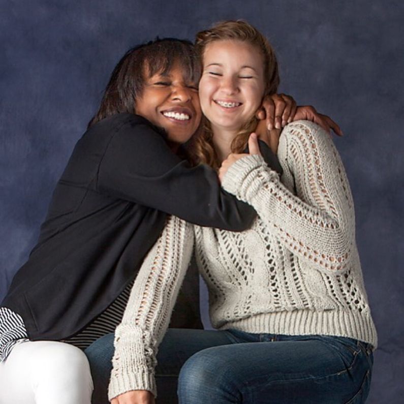 Two smiling teenage girls embrace each other in a studio portrait with blue-gray background.
