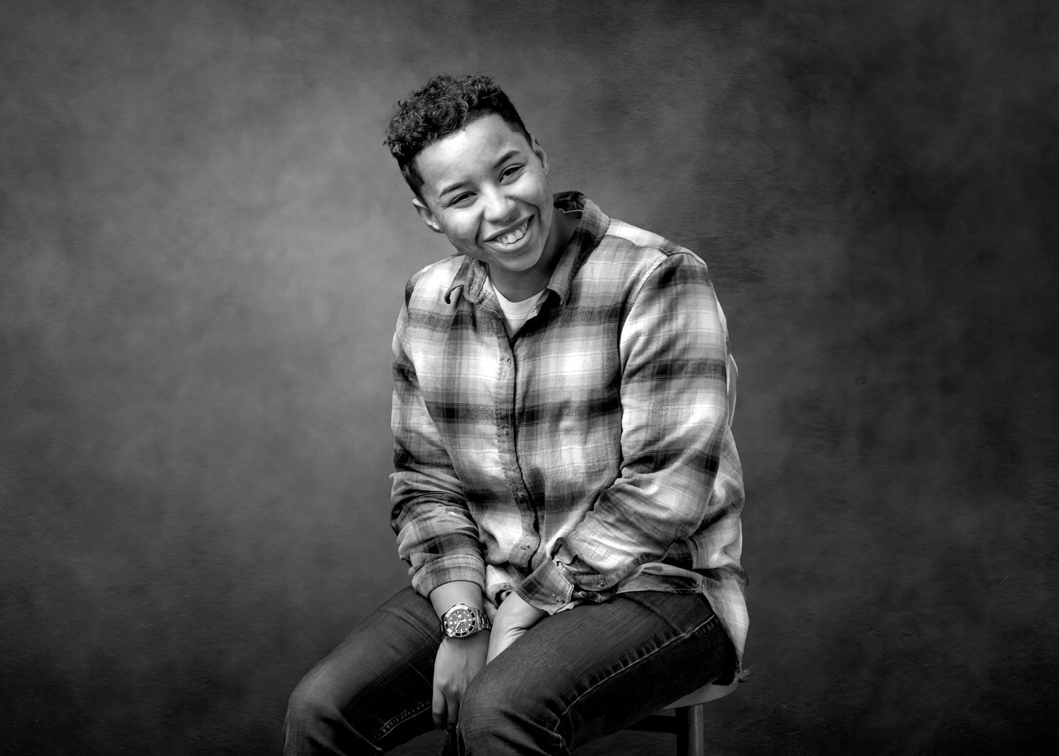 Black and white portrait of smiling person in plaid shirt seated on stool.