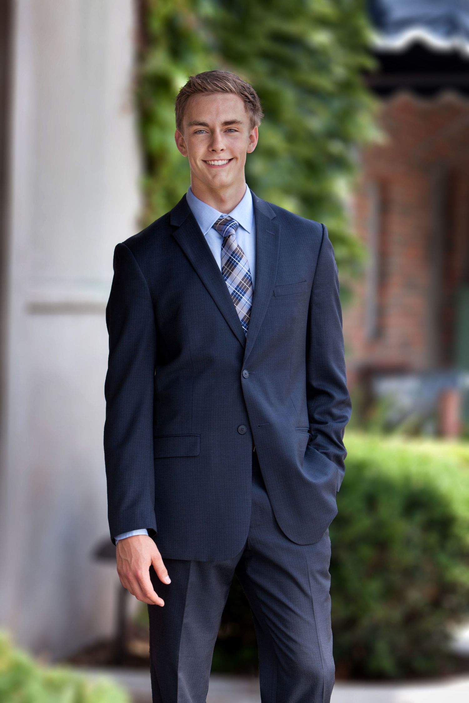 Senior boy in navy suit and plaid tie smiling outdoors near brick building, by Jocelyn M. Photography in Rochester, MI