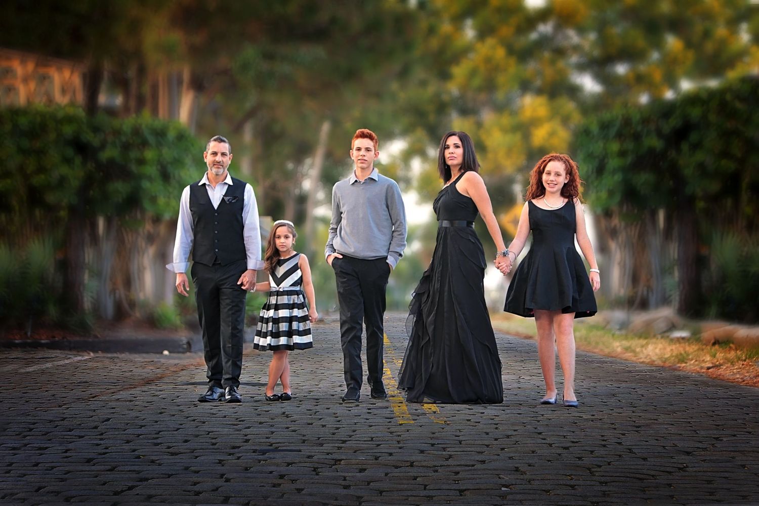 Family of five posing on a tree-lined path, dressed in elegant black and white outfits for a formal portrait.