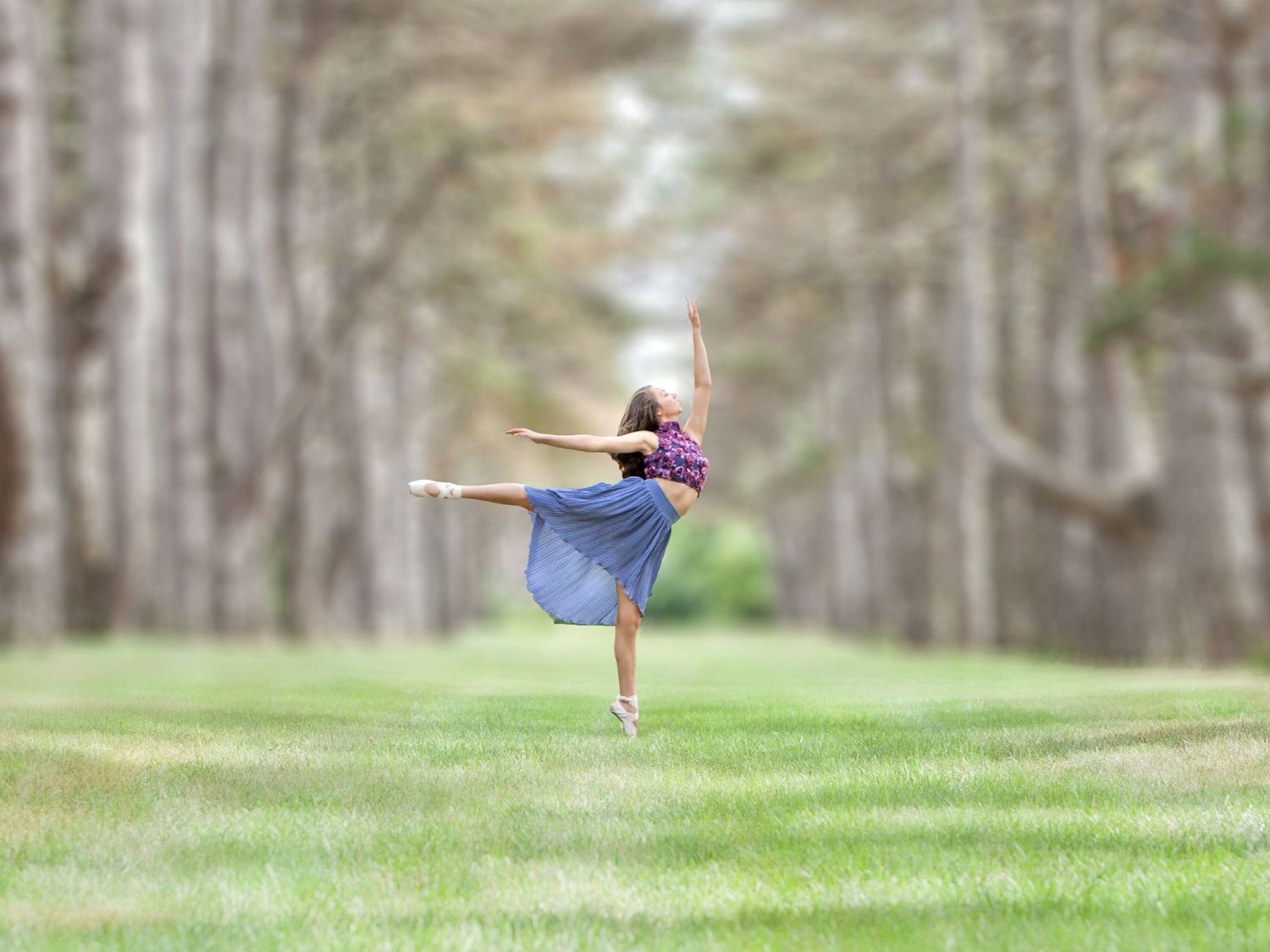 Graceful dancer in blue skirt performing a ballet pose in a lush green tree-lined field.