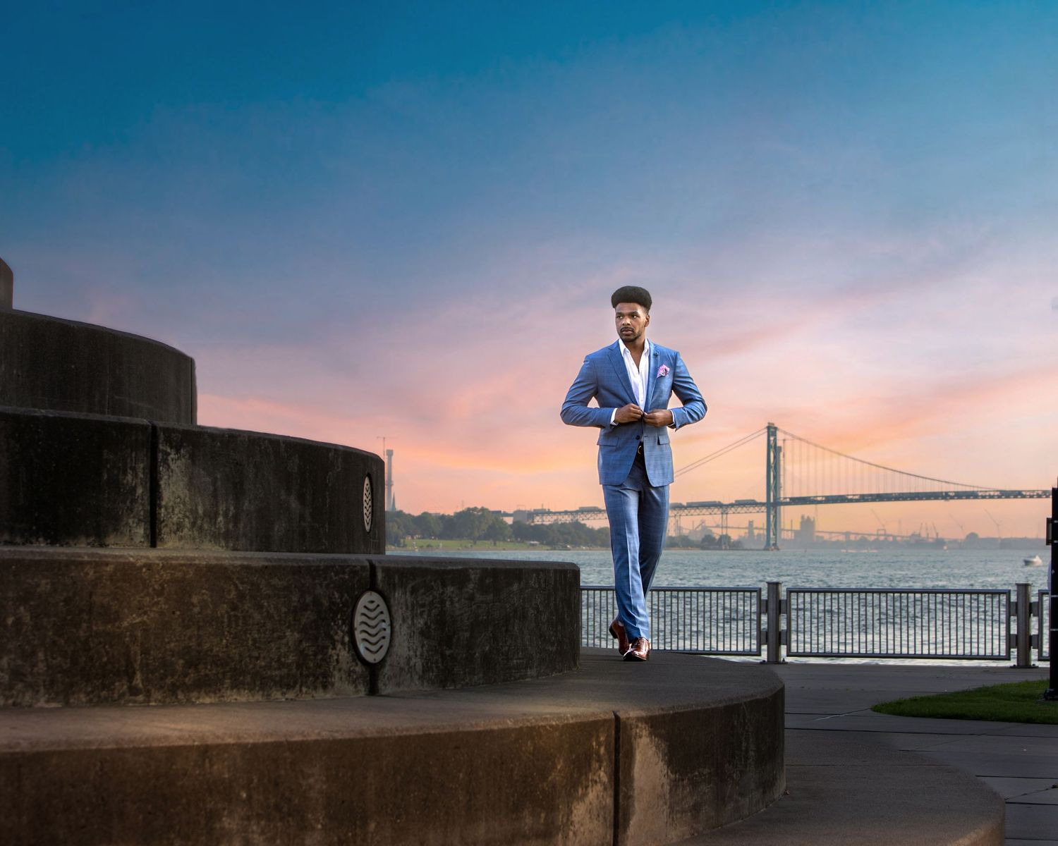 Man in blue suit walks confidently near waterfront with Golden Gate Bridge at sunset behind him.