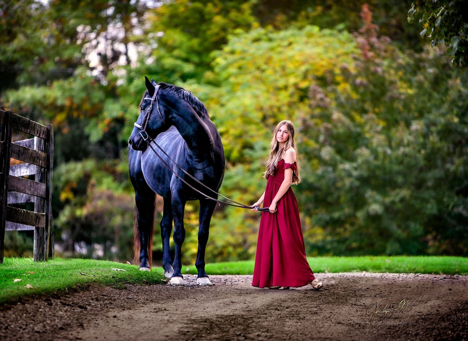 Young woman in red gown leads a dark horse along a scenic path surrounded by lush green trees.