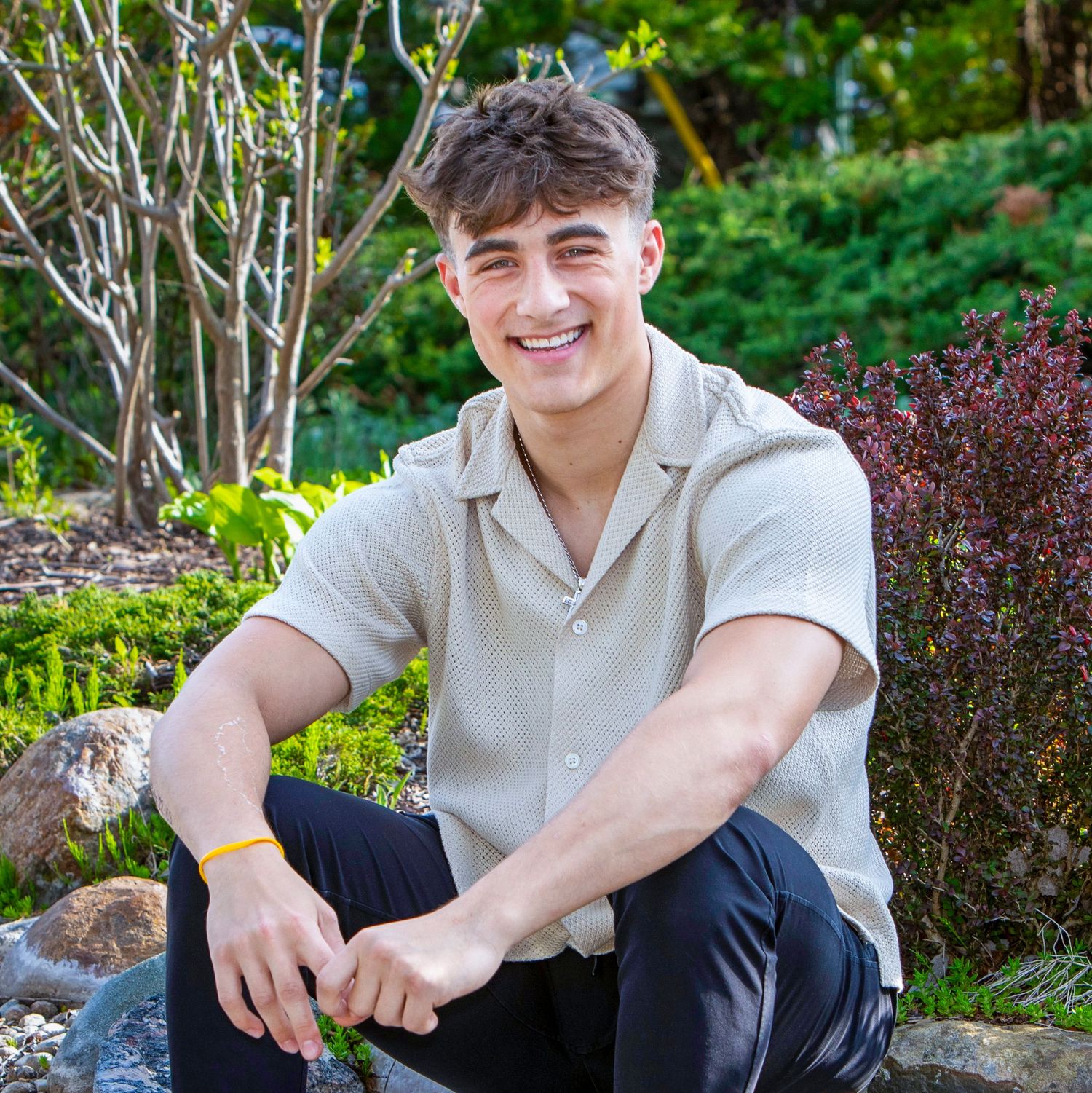 Smiling young man in a light button-up shirt sitting outdoors in a lush green garden setting.