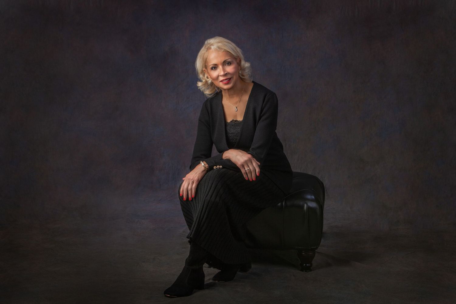 Elegant woman in black outfit sits gracefully on ottoman for professional studio portrait.