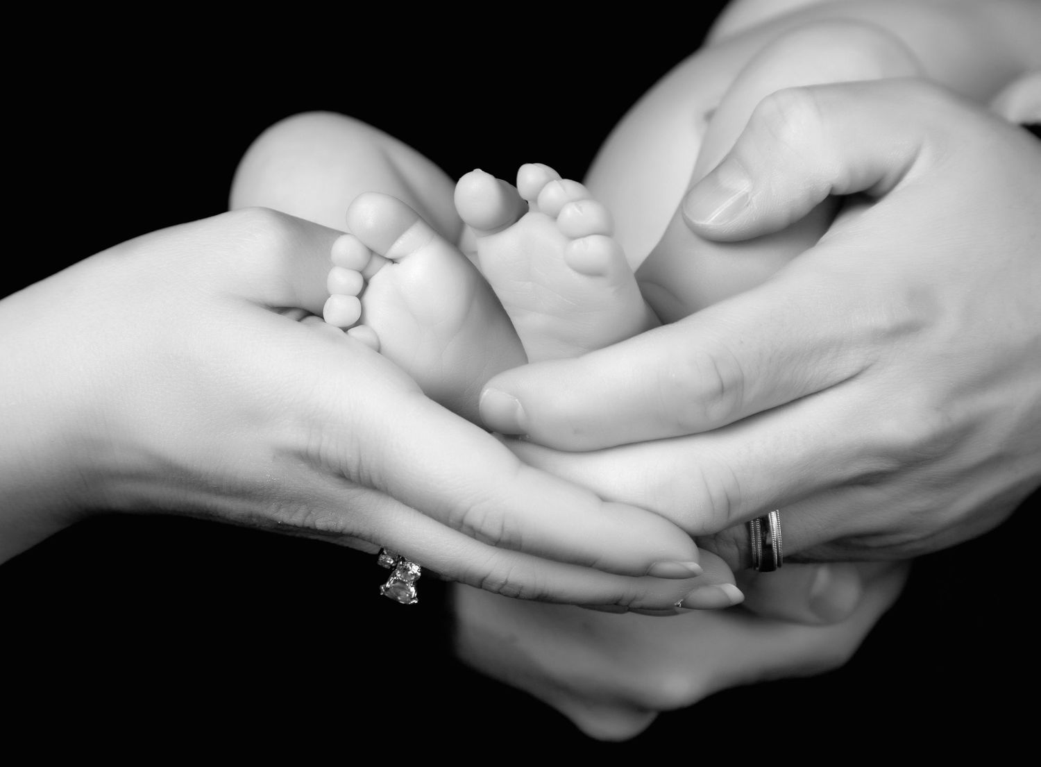 Black and white photo of parents cradling newborn baby feet, mother wearing diamond ring.