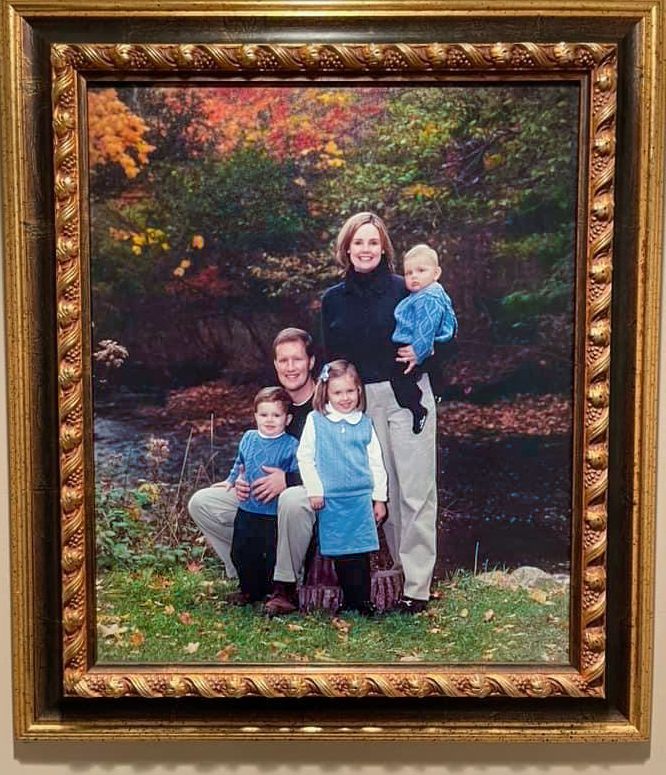 Framed family portrait with parents and three young children posed outdoors among colorful autumn foliage.