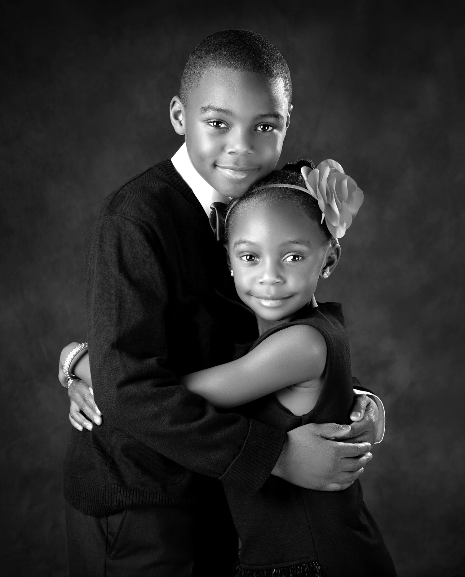 Black and white portrait of two children hugging, a boy in a suit and a girl with a bow, smiling warmly.