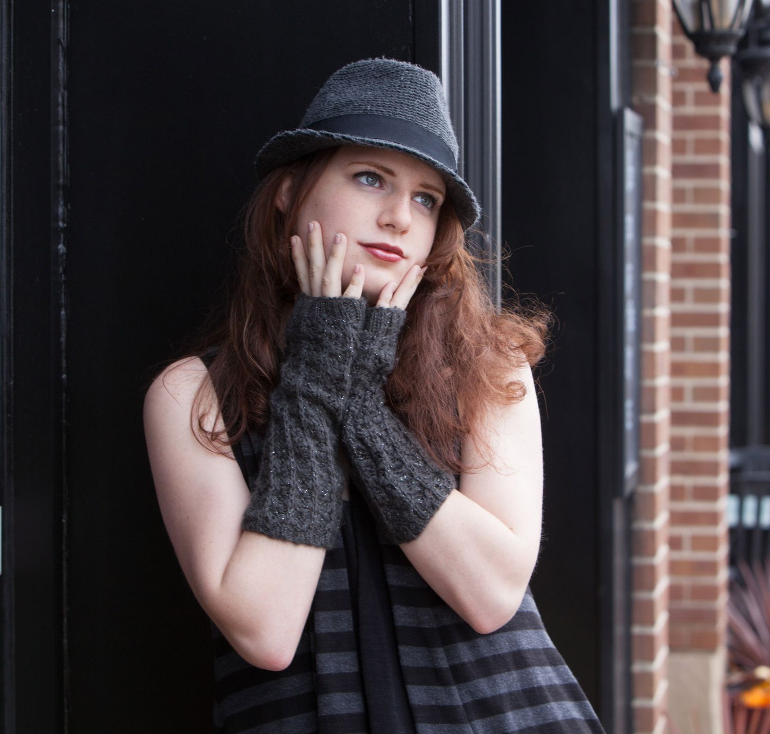 Stylish redhead woman in a gray hat and fingerless gloves posing thoughtfully by a dark door.
