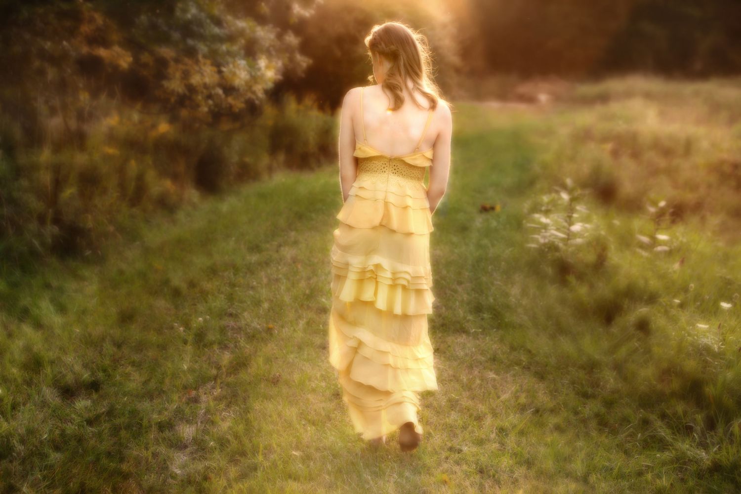 Woman in yellow ruffled maxi dress walking along a sunlit green path surrounded by lush trees at golden hour.
