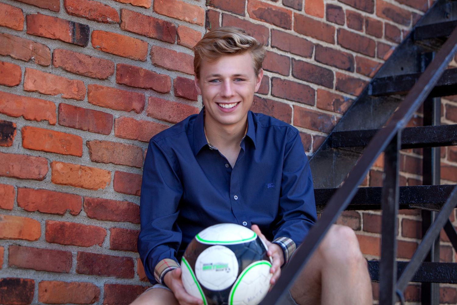 Smiling teenage boy holding a soccer ball while sitting on steps against a red brick wall.