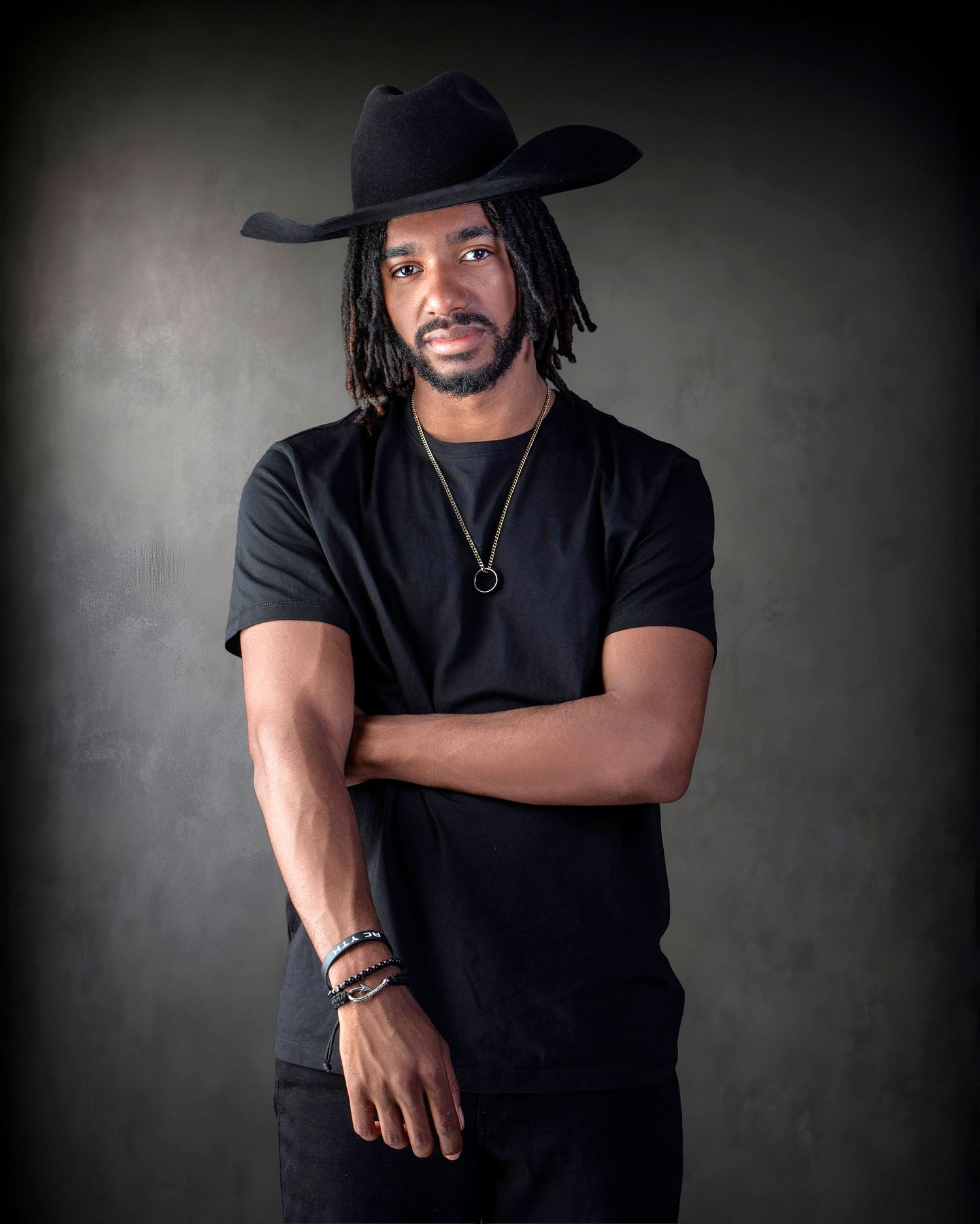 Stylish man in black outfit and cowboy hat with dreadlocks posing with arms crossed in studio.