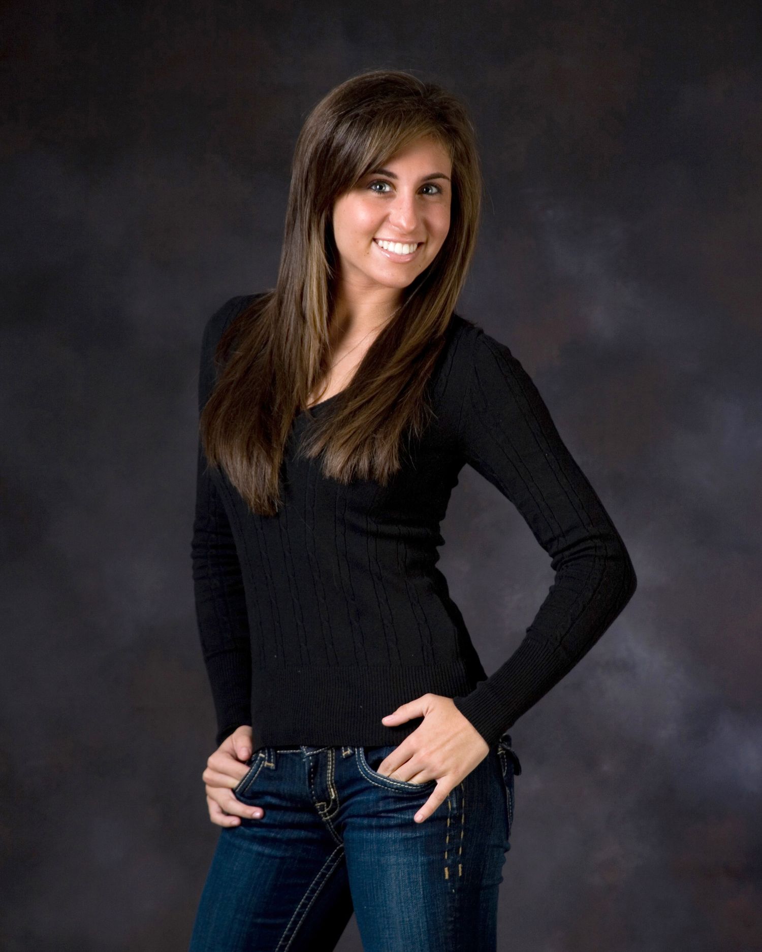 Smiling young woman with long brown hair wearing a black sweater and jeans in a studio portrait.