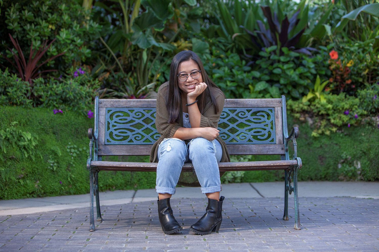 Young woman sitting on a park bench smiling, surrounded by lush green garden foliage.