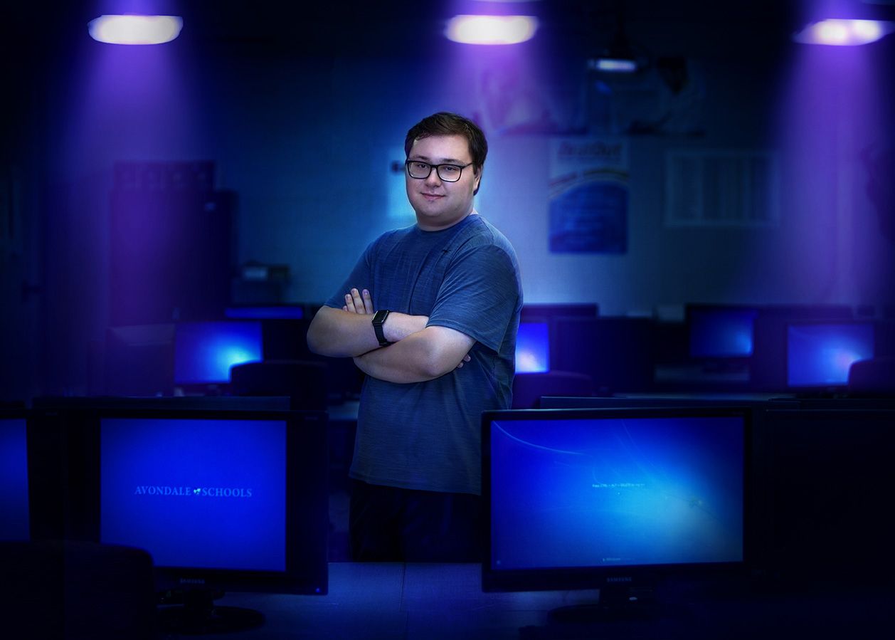 Young man standing confidently with arms crossed between glowing computer monitors in blue-lit room.