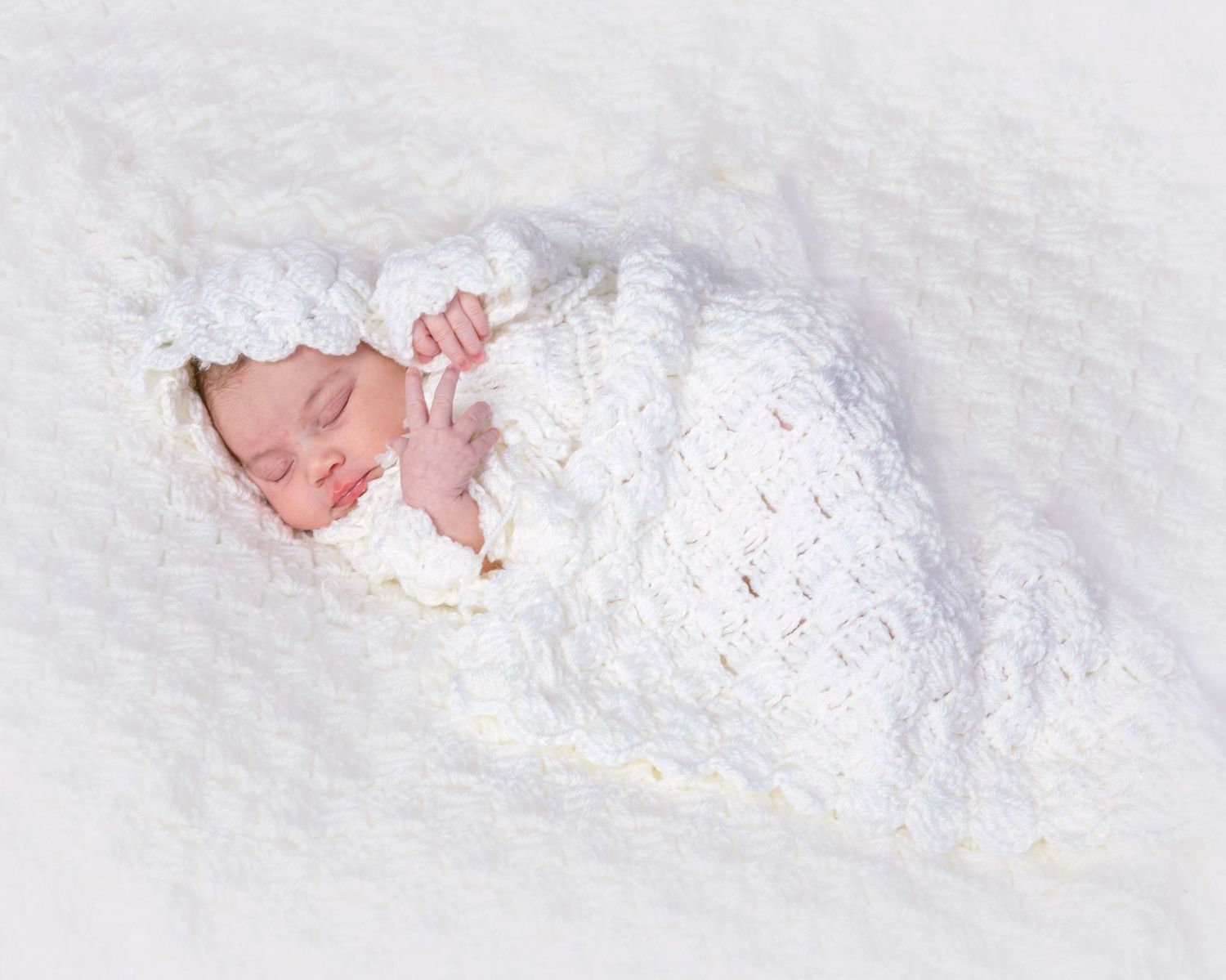 Sleeping newborn baby wrapped in a white crocheted cocoon swaddle with matching hat on a white background.