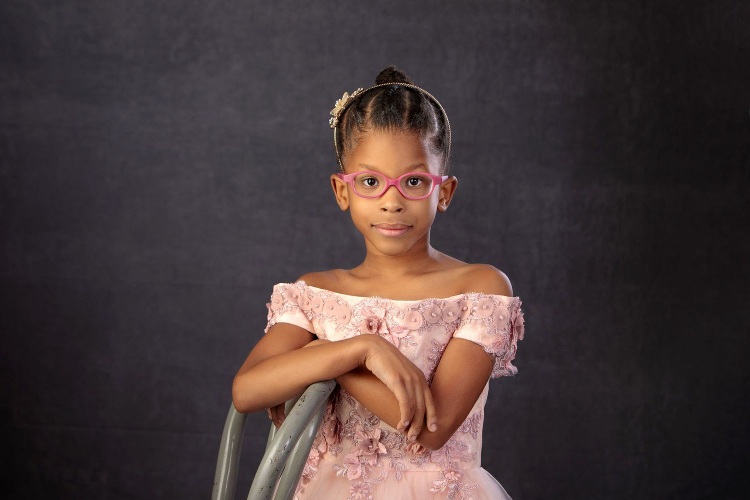 Young Black girl in pink off-shoulder dress and pink glasses poses confidently against dark background.