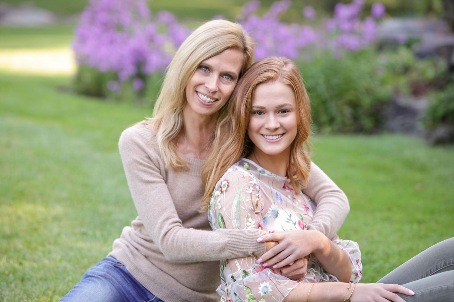 Two smiling women pose together outdoors on green grass with purple flowers in the background.