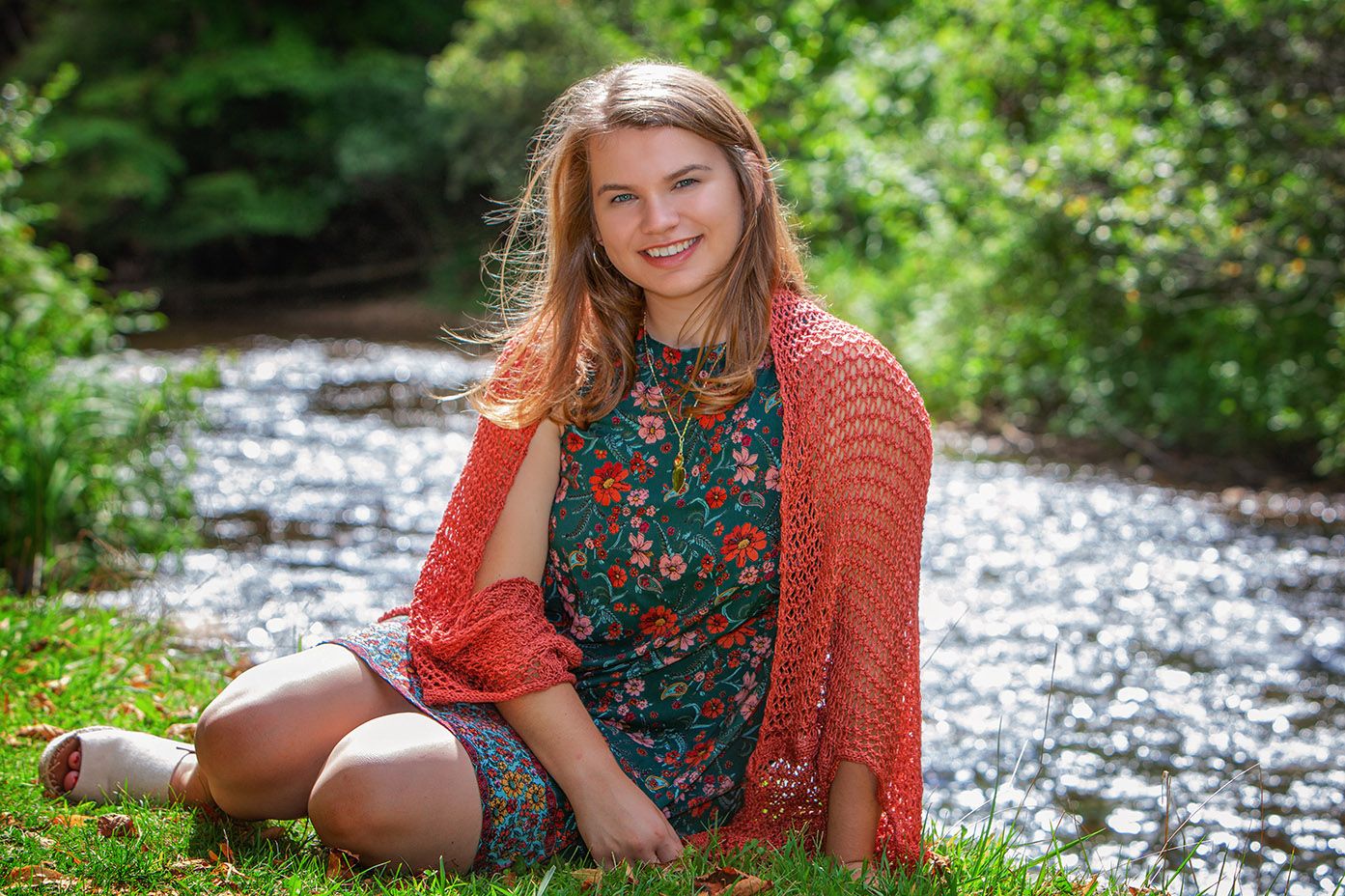 Smiling young girl in floral dress with orange shawl sitting on grass beside a sunlit forest stream.