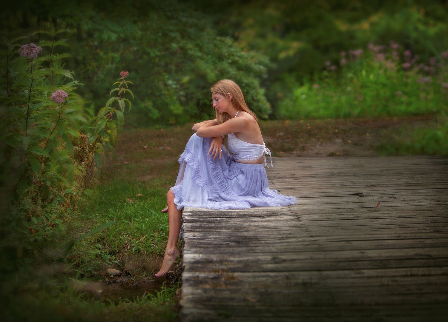 Young girl in a light blue dress sitting pensively on a wooden dock surrounded by lush greenery.