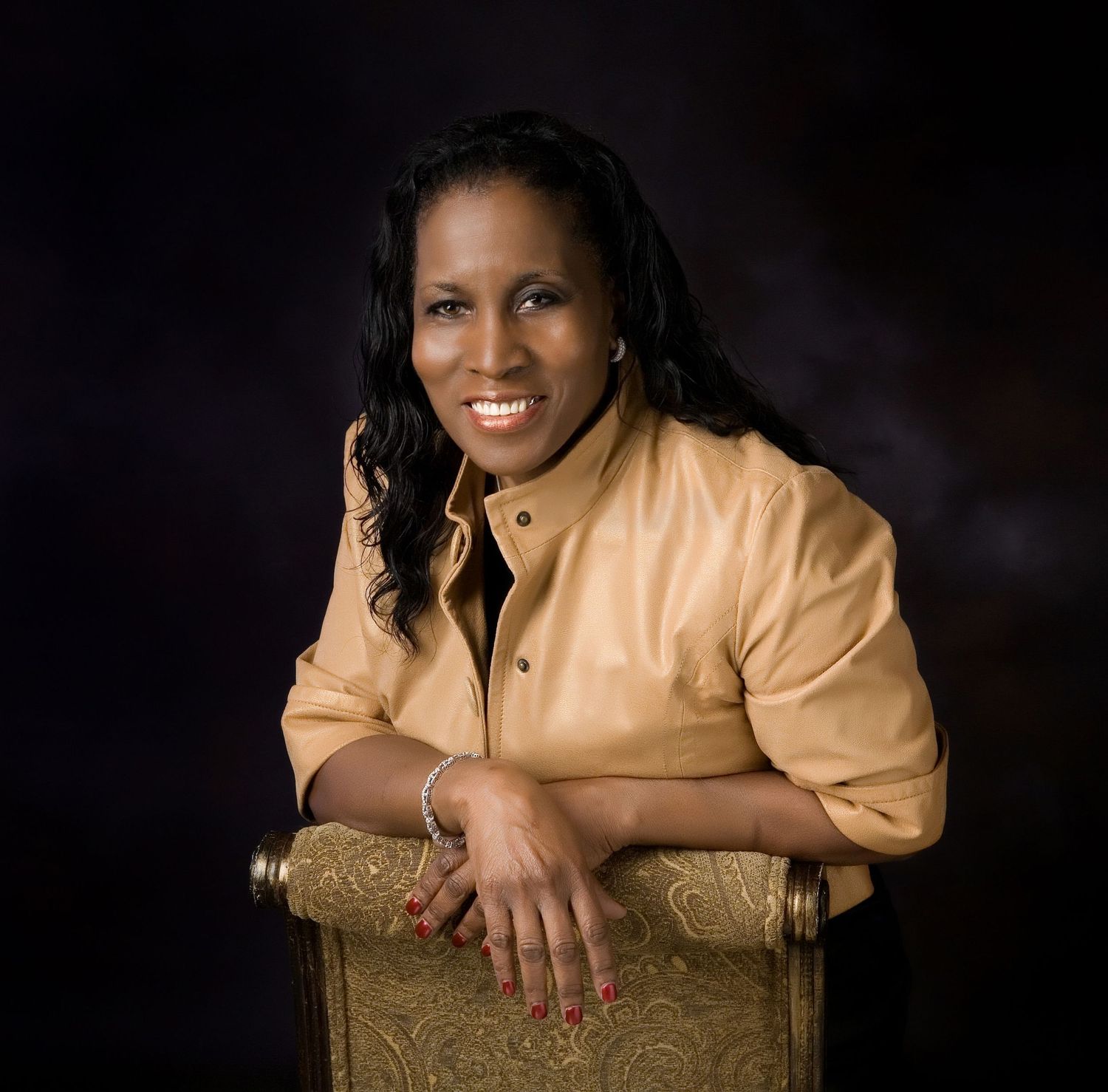 Confident woman in gold jacket smiling while seated, posed against dark studio background.