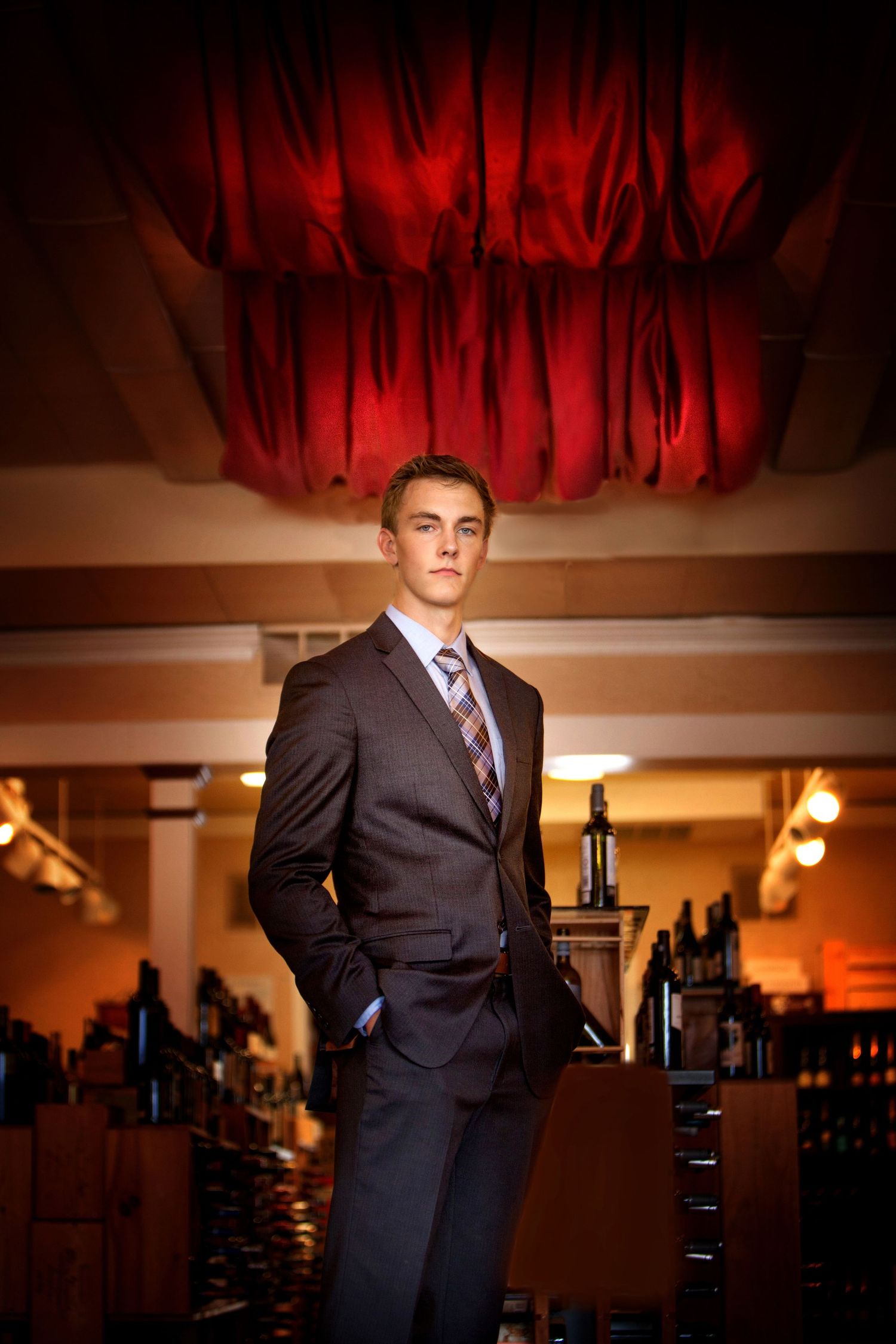 Senior boy in dark suit and tie stands confidently in an upscale bar with red draped ceiling, by Jocelyn M. Photography in Rochester, MI
