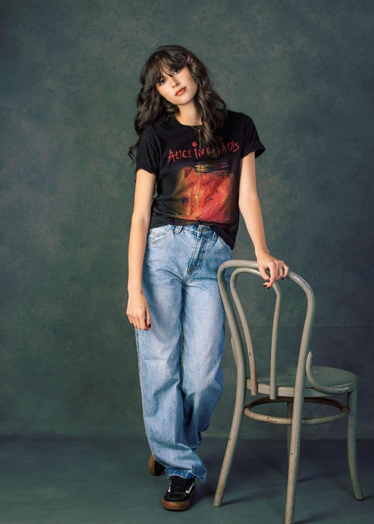 Young woman in Alice in Chains tee and wide-leg jeans leans on chair in studio.