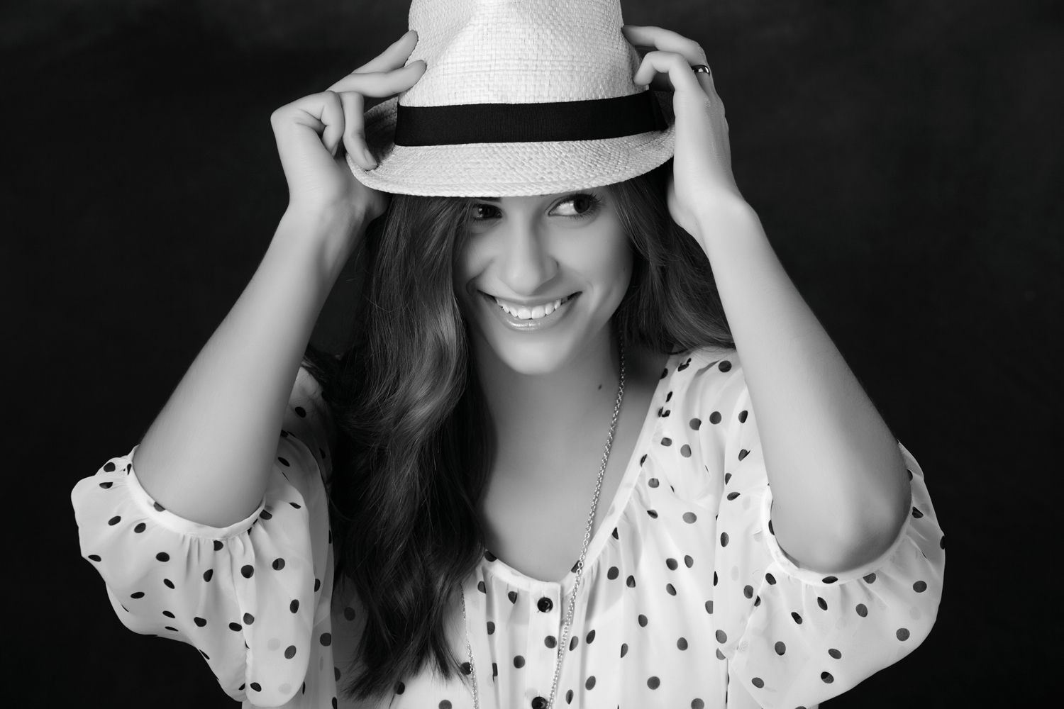 Smiling brunette woman holding a fedora hat in a black and white portrait.