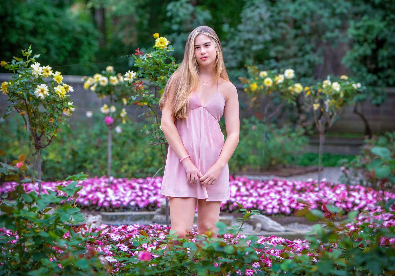 Young woman in pink dress standing among colorful blooming flowers in a lush garden park.