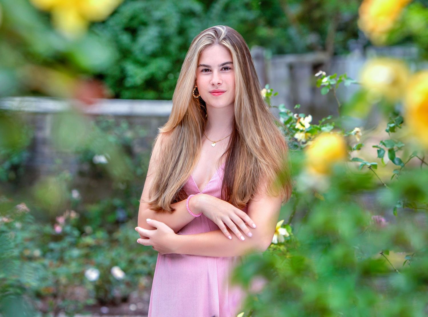Young woman with long blonde hair in a pink dress posing gracefully among lush green foliage and yellow roses.