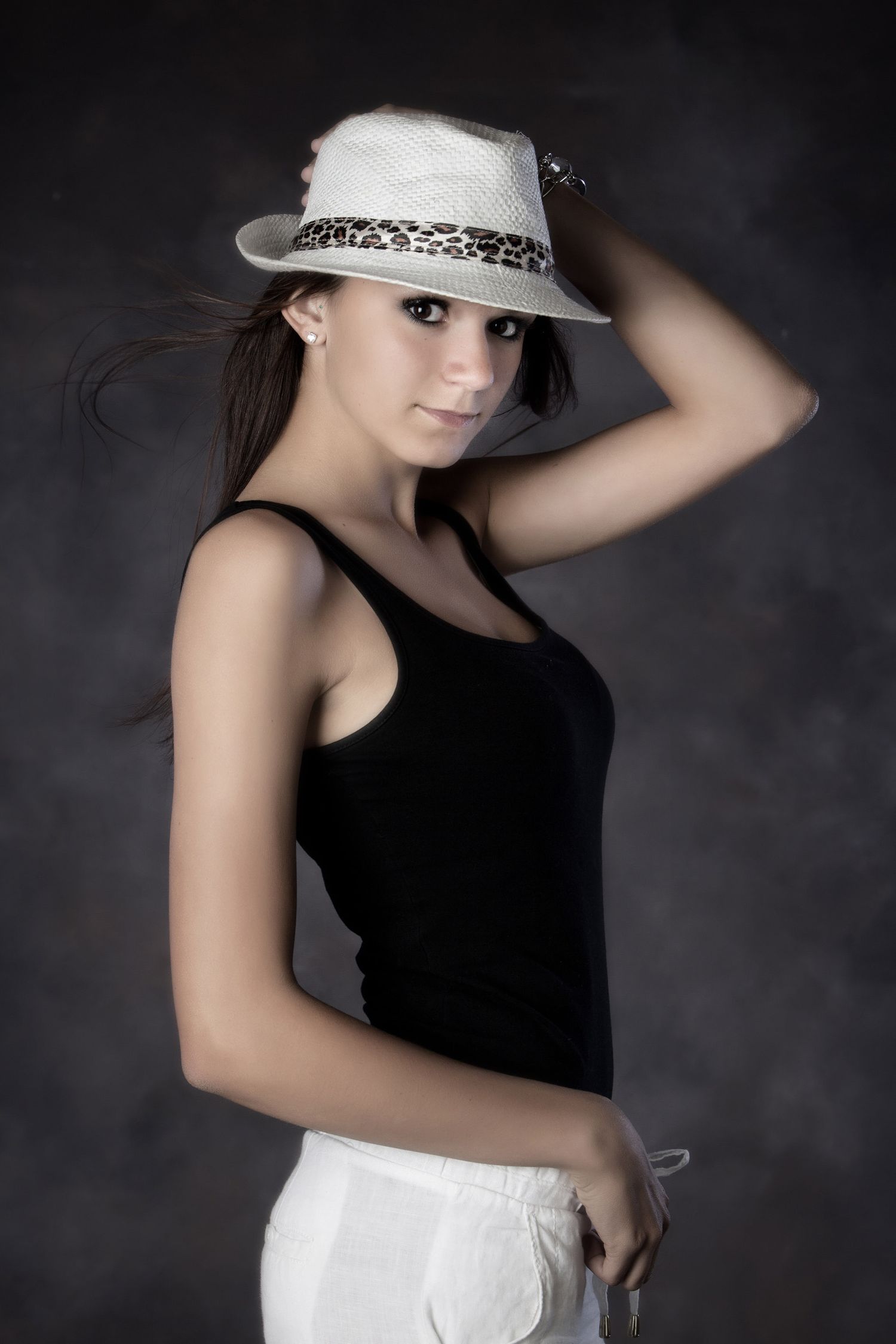 Young woman in black tank top and white fedora hat with leopard band posing confidently against dark background.
