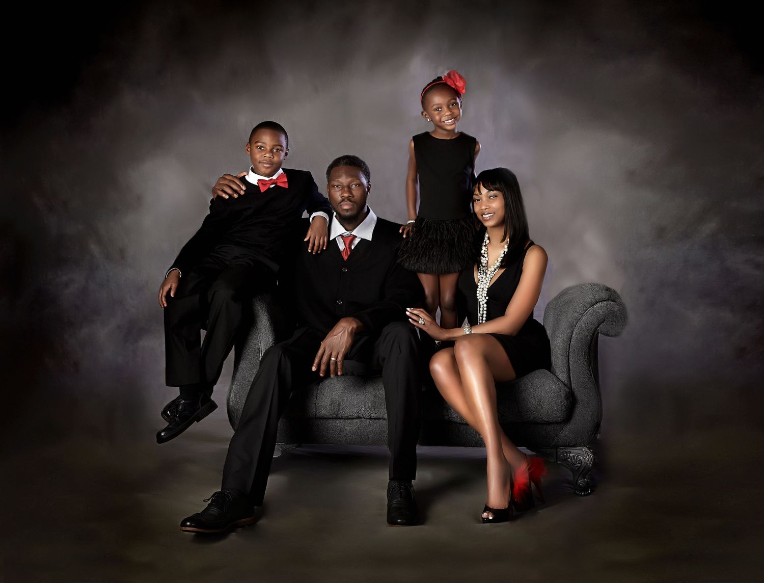Elegant family portrait of four in formal black and red attire posed on a gray velvet chair against dark background.