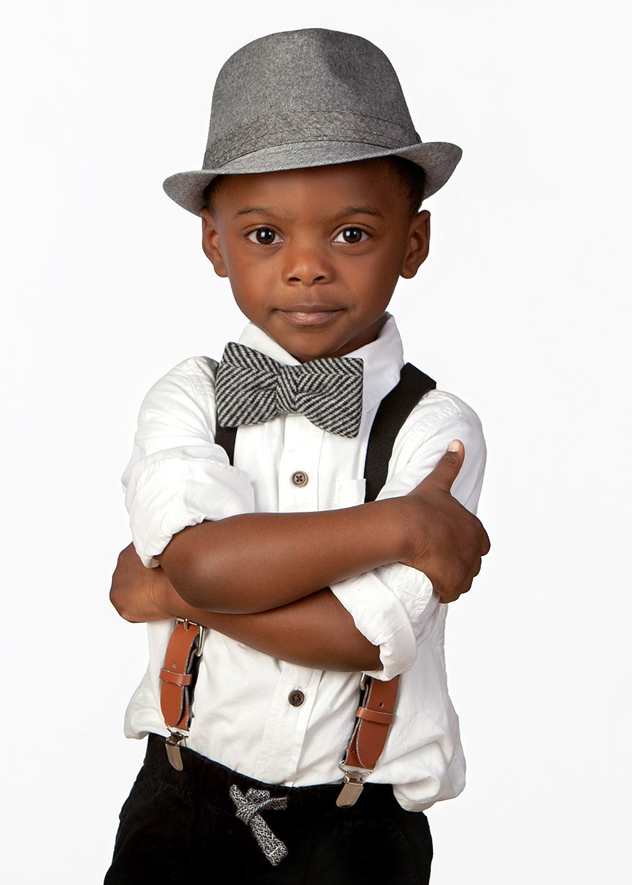 Stylish young boy in gray fedora, bow tie, suspenders, and white shirt posing confidently with arms crossed.