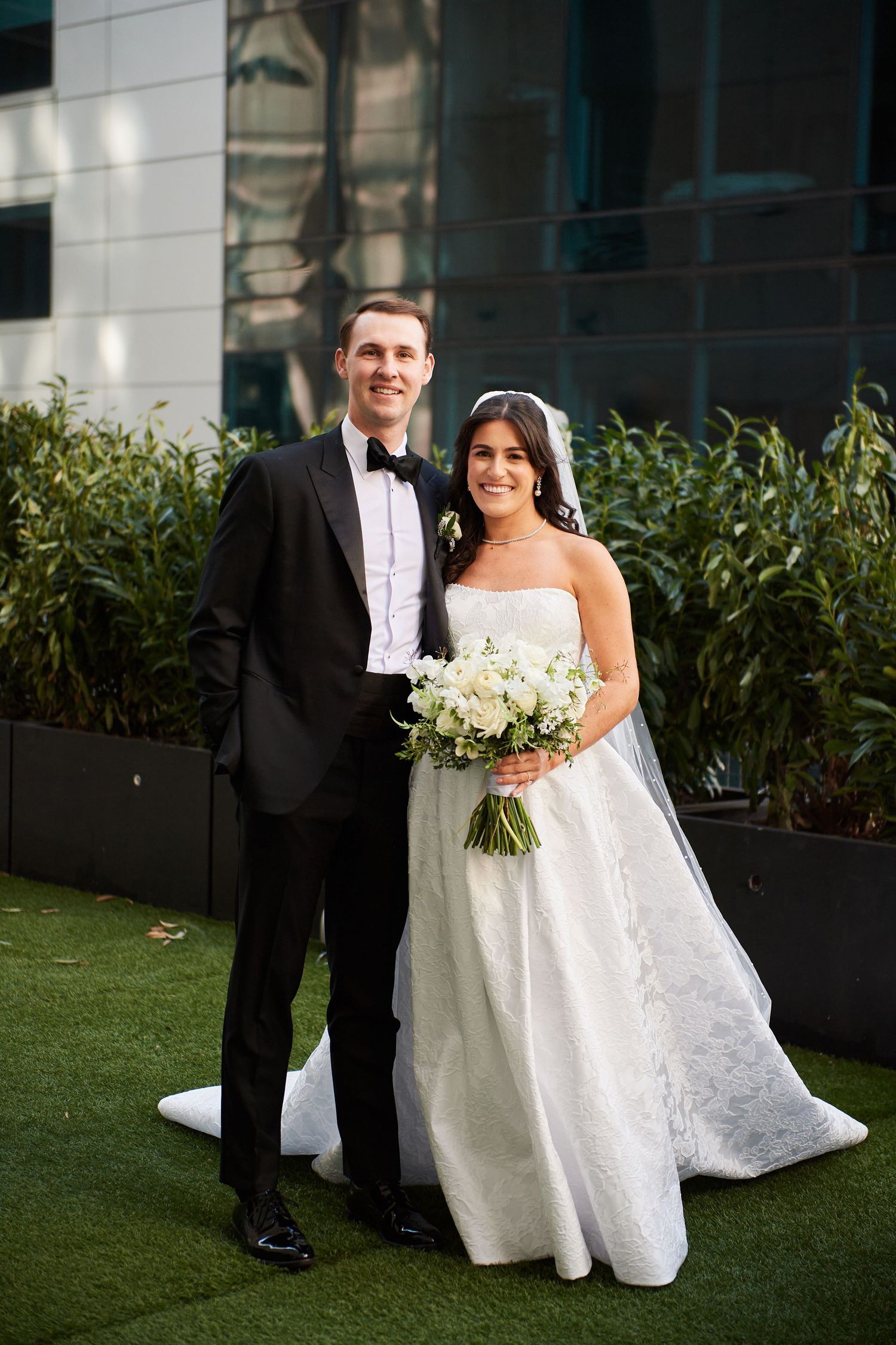Bride and groom posing outdoors on green grass, bride in white strapless gown holding bouquet, groom in black tuxedo.