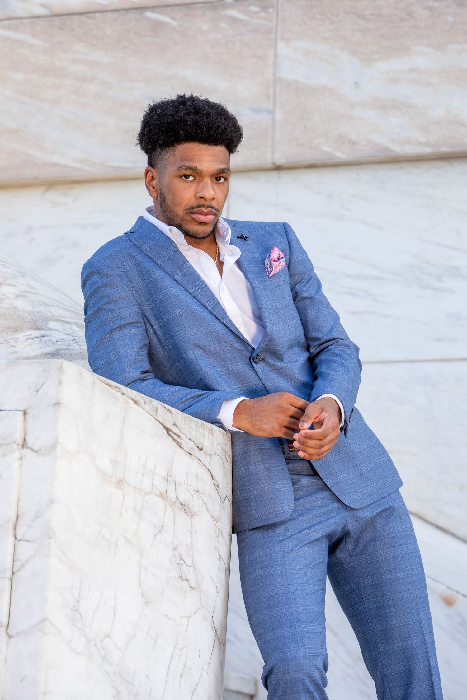 Young Black man in blue suit with pink pocket square leans against marble pillar outdoors.