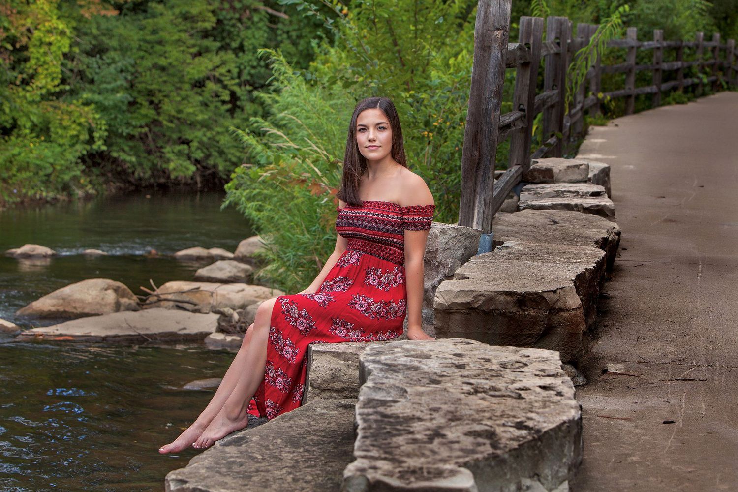 Young woman in red floral off-shoulder dress sitting on stone wall by a peaceful creek surrounded by lush greenery.