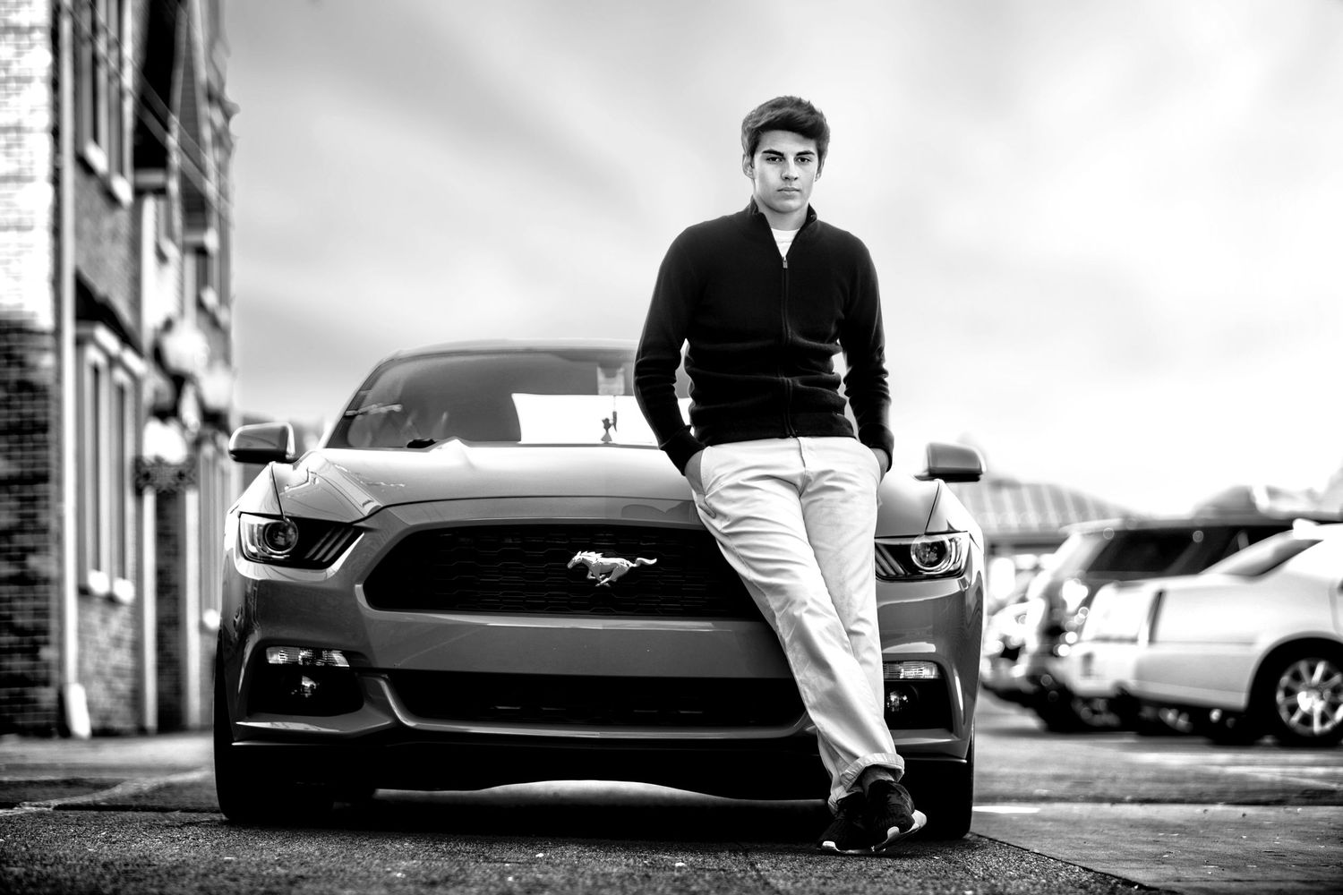 Young man in black sweater poses confidently beside a Ford Mustang in a stylish black and white outdoor photo.