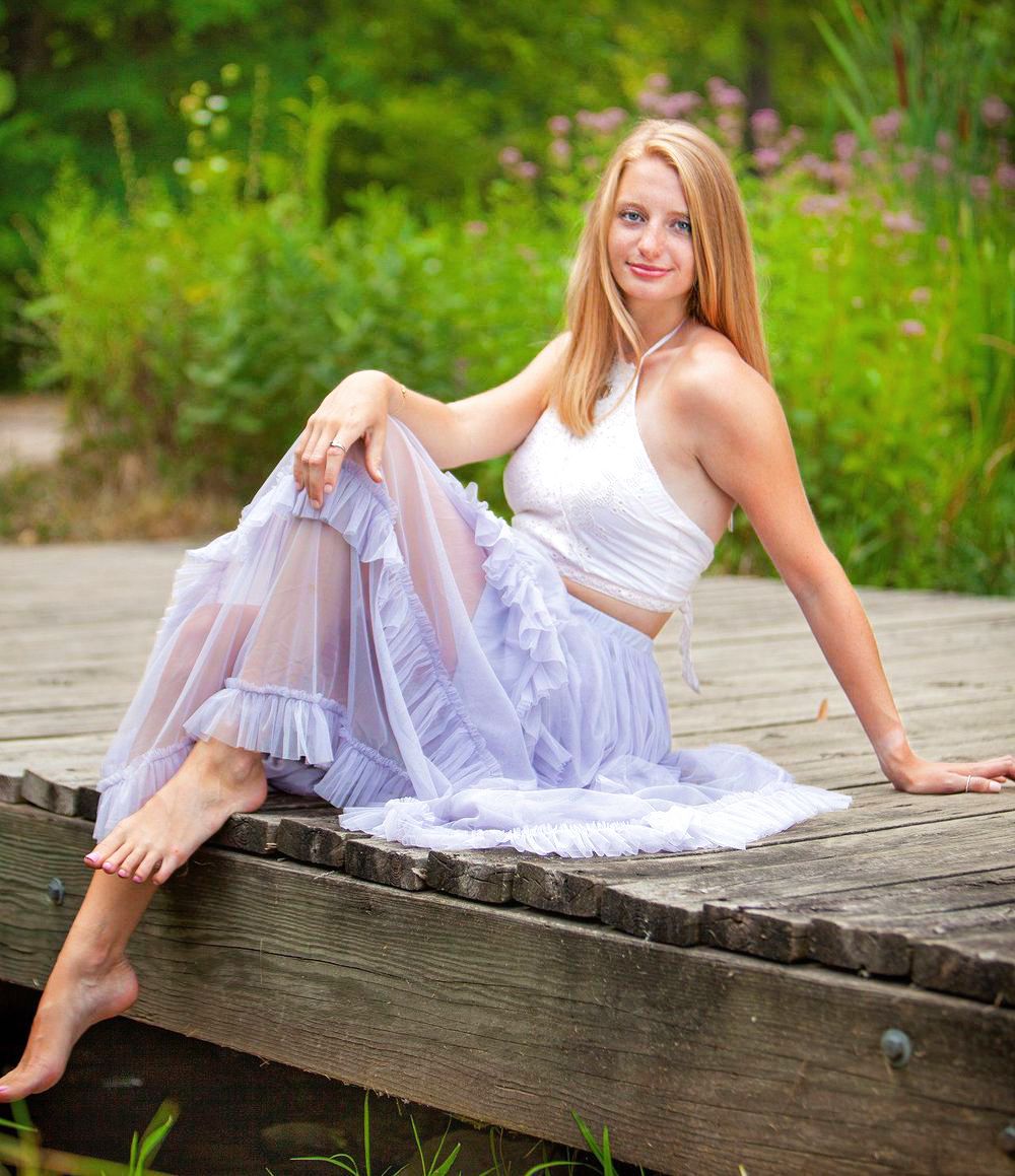 Senior girl in white top and lavender ruffle skirt sitting barefoot on wooden deck outdoors. by Jocelyn M. Photography in Rochester, MI