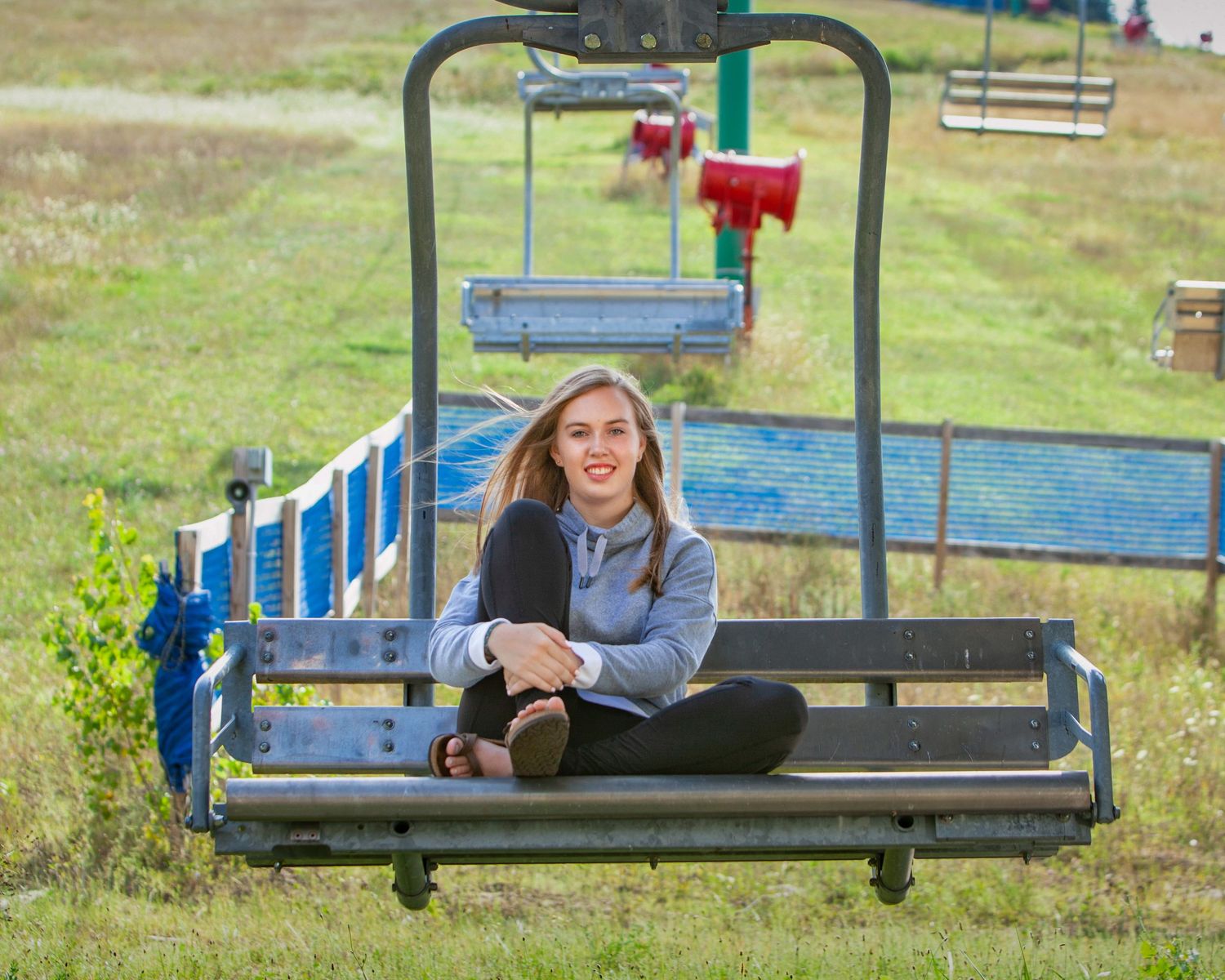 Smiling teenage girl sitting on a ski lift chair on a grassy hillside on a sunny day.