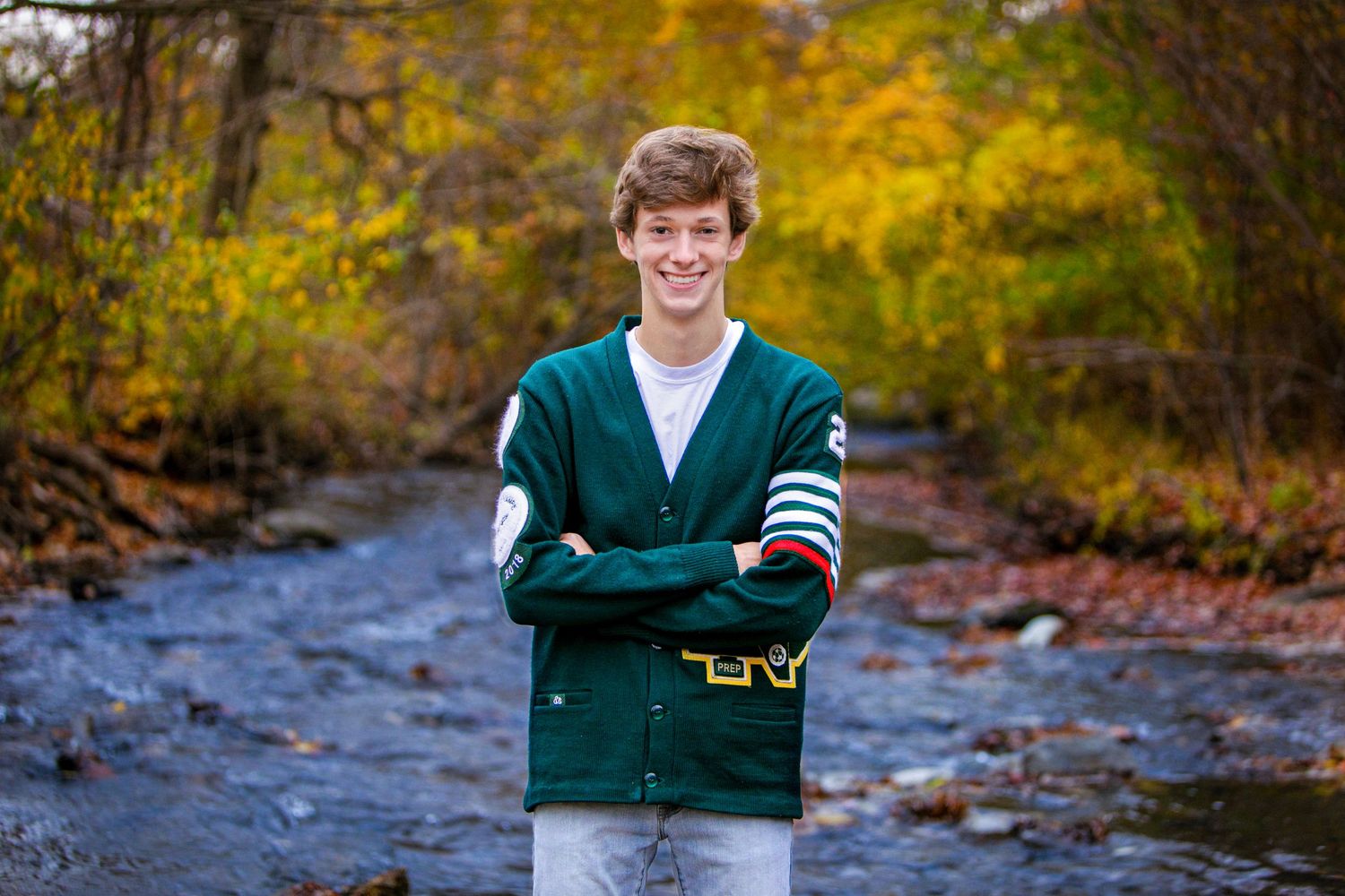 Smiling teen boy in green varsity cardigan standing by a stream surrounded by fall foliage.