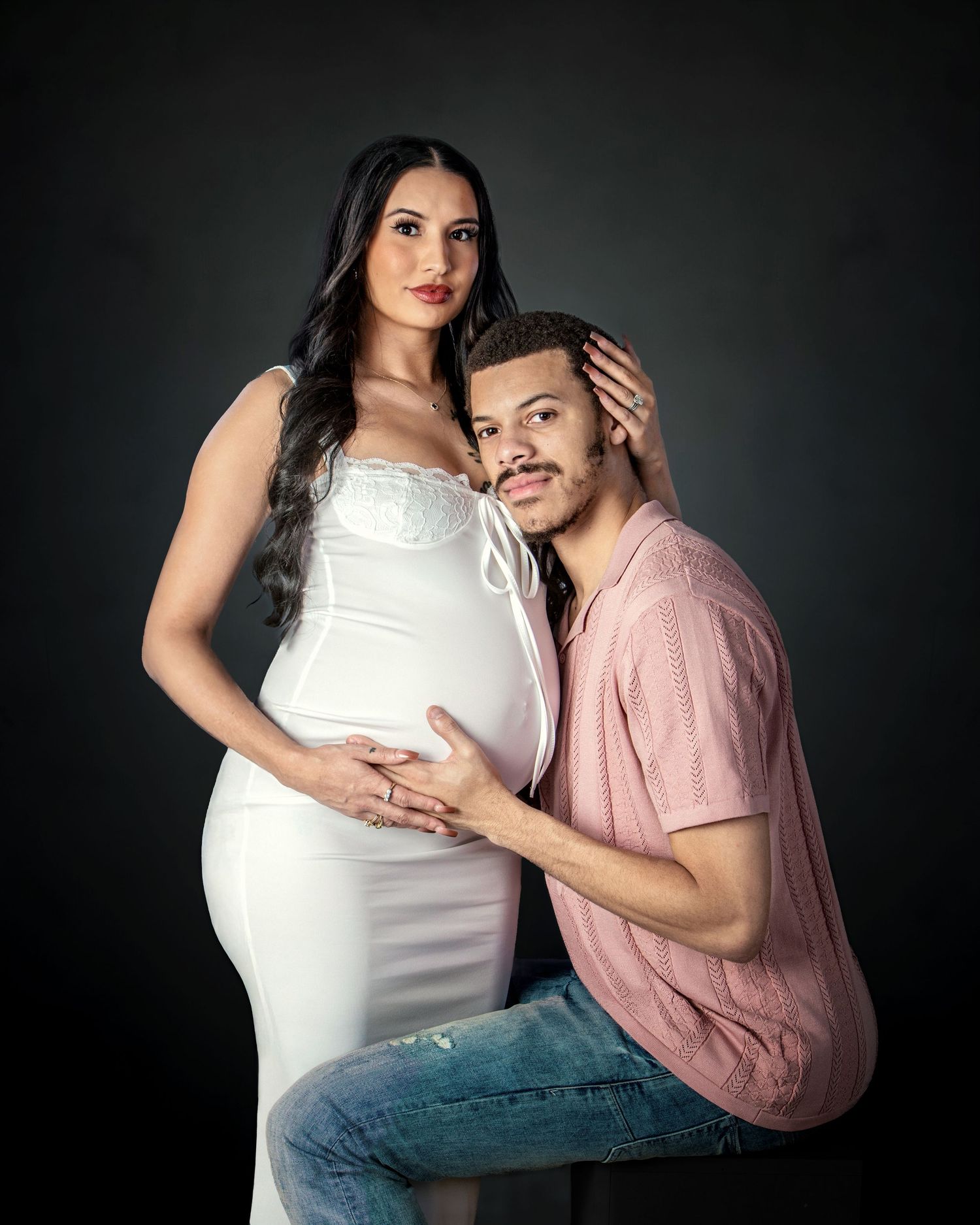 Couple poses for maternity photo shoot, pregnant woman in white dress with partner in pink shirt on dark background.