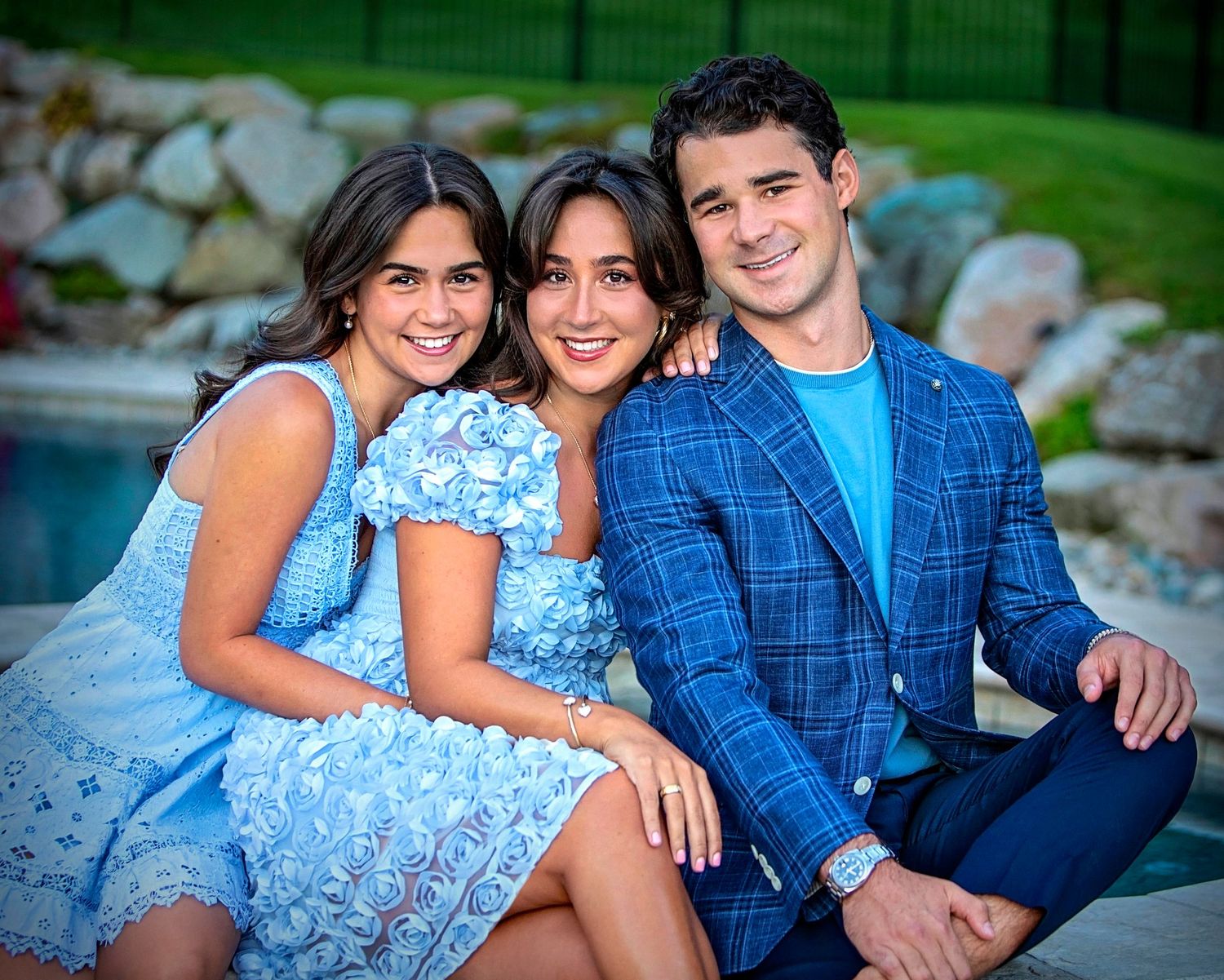 Three smiling people in coordinating blue outfits pose together outdoors near a pool on a sunny day.