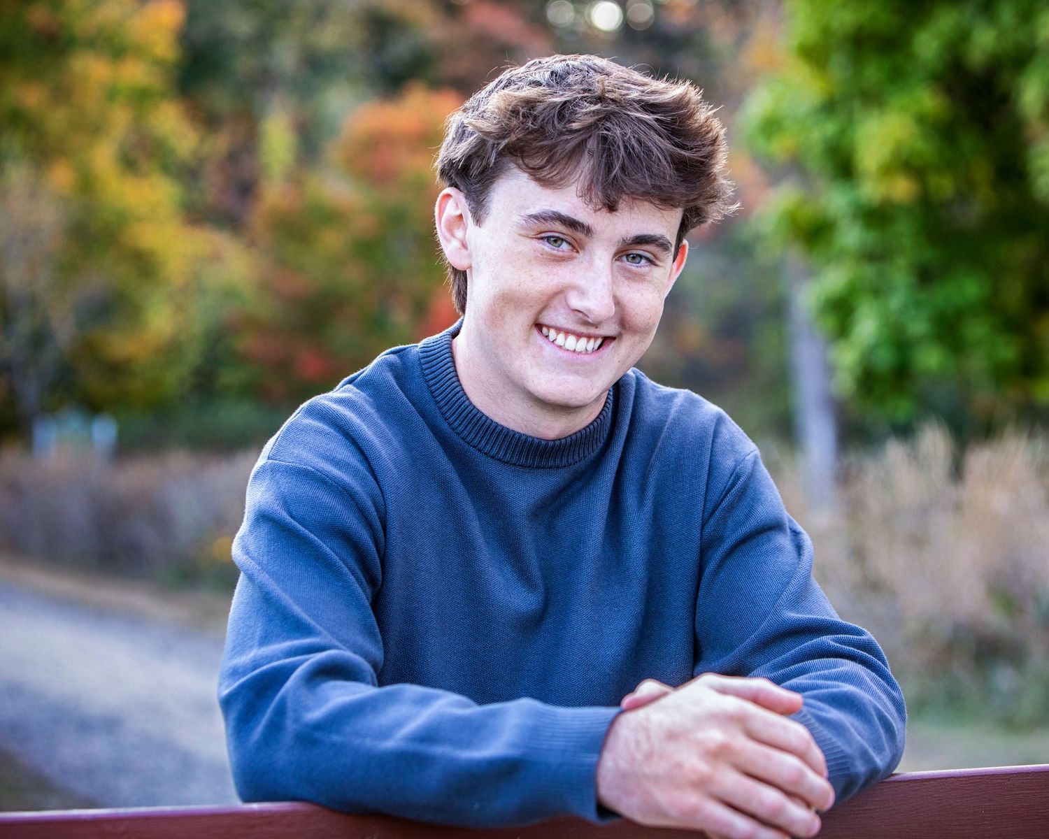 Smiling young man in blue sweater leaning on a red fence outdoors with colorful autumn foliage background.