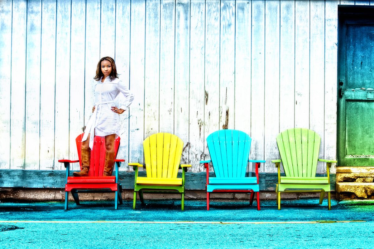 Woman in white dress standing beside colorful red, yellow, and green Adirondack chairs against a rustic wall.