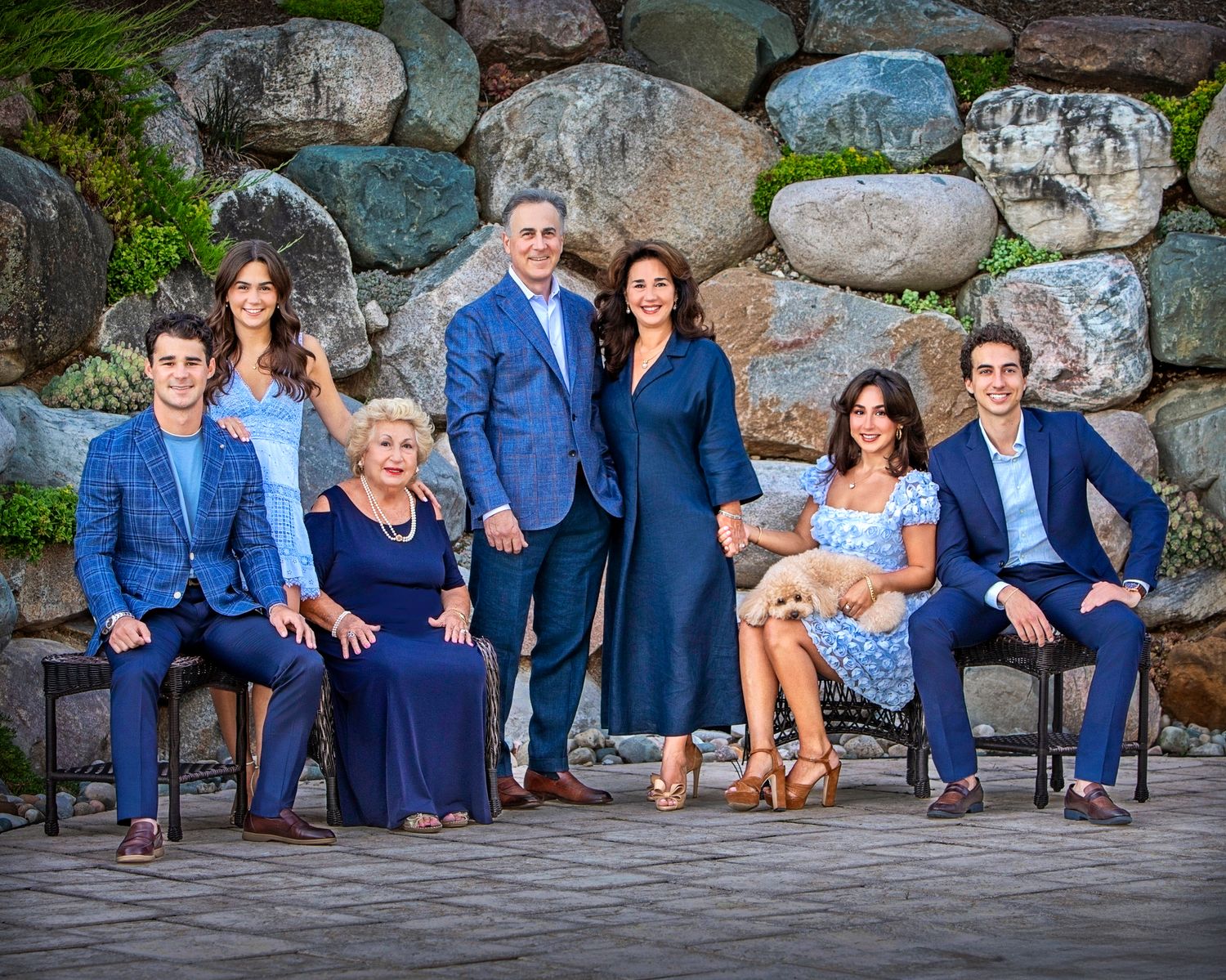 A multigenerational family poses outdoors in coordinating blue outfits with a golden puppy, set against a stone wall backdrop.